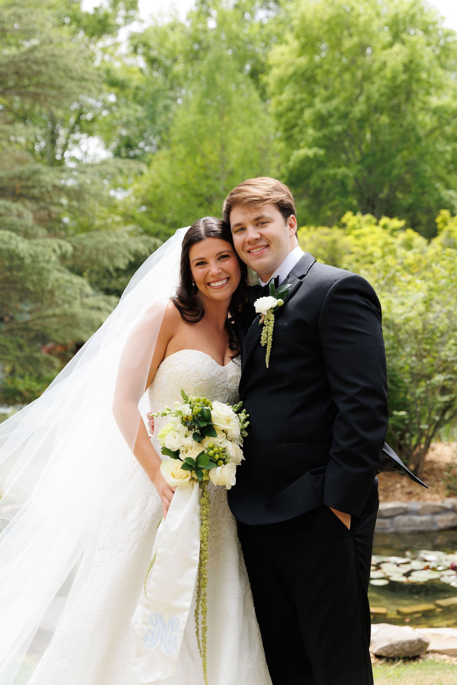 Smiling bride and groom pose outdoors in garden setting, bride in strapless gown with veil holding white bouquet.