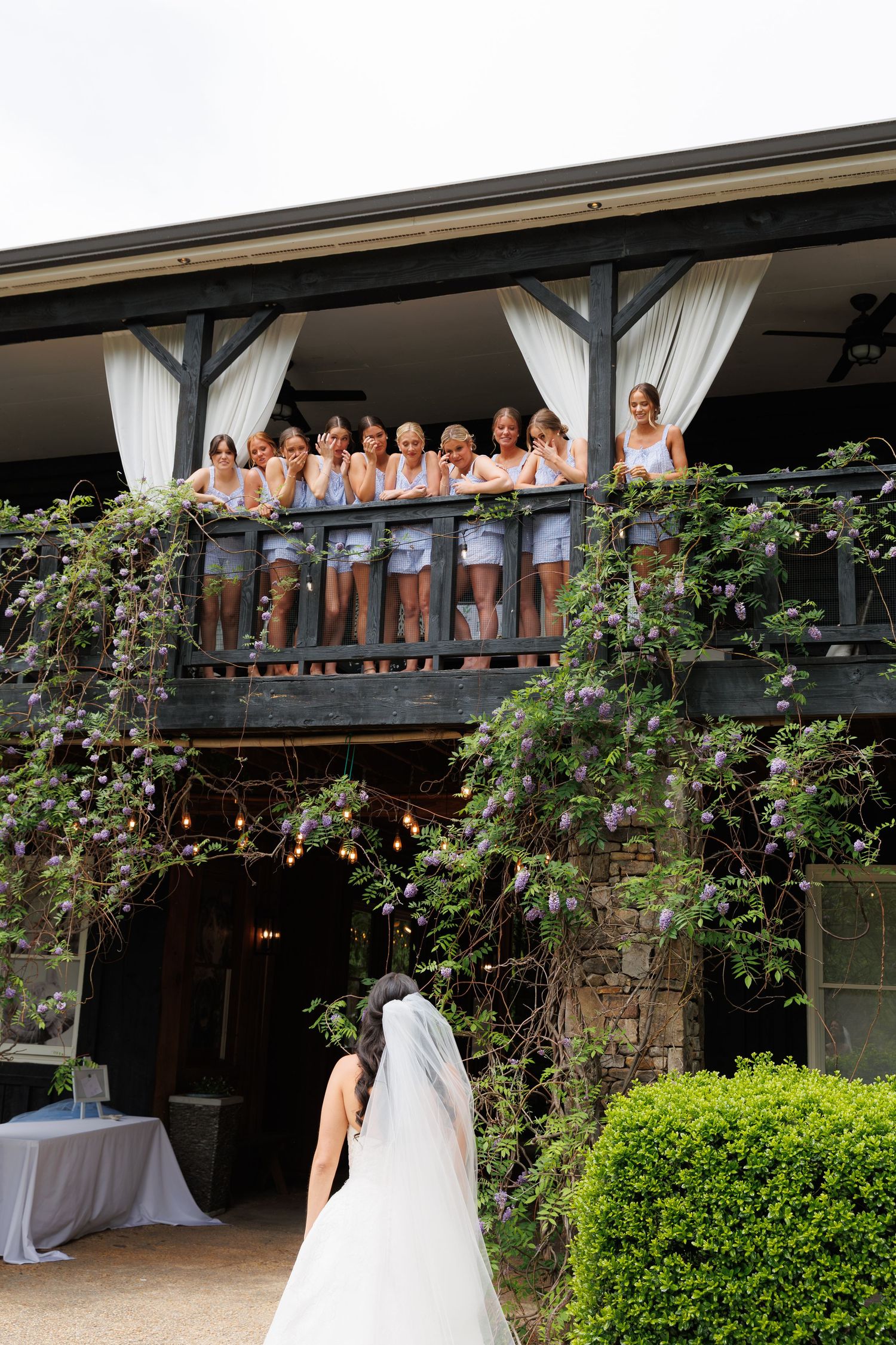 Bridesmaids lean over a rustic balcony adorned with wisteria, reacting with joy as the bride reveals her wedding dress below.
