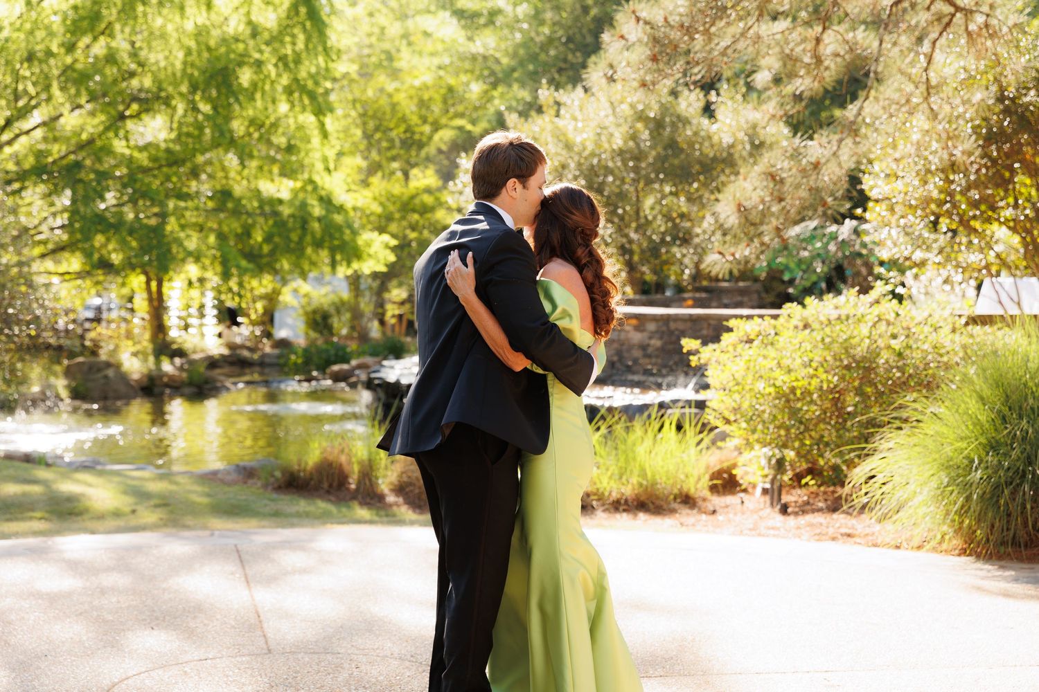 Couple sharing a romantic kiss in a sunlit garden, woman in green gown and man in dark suit.