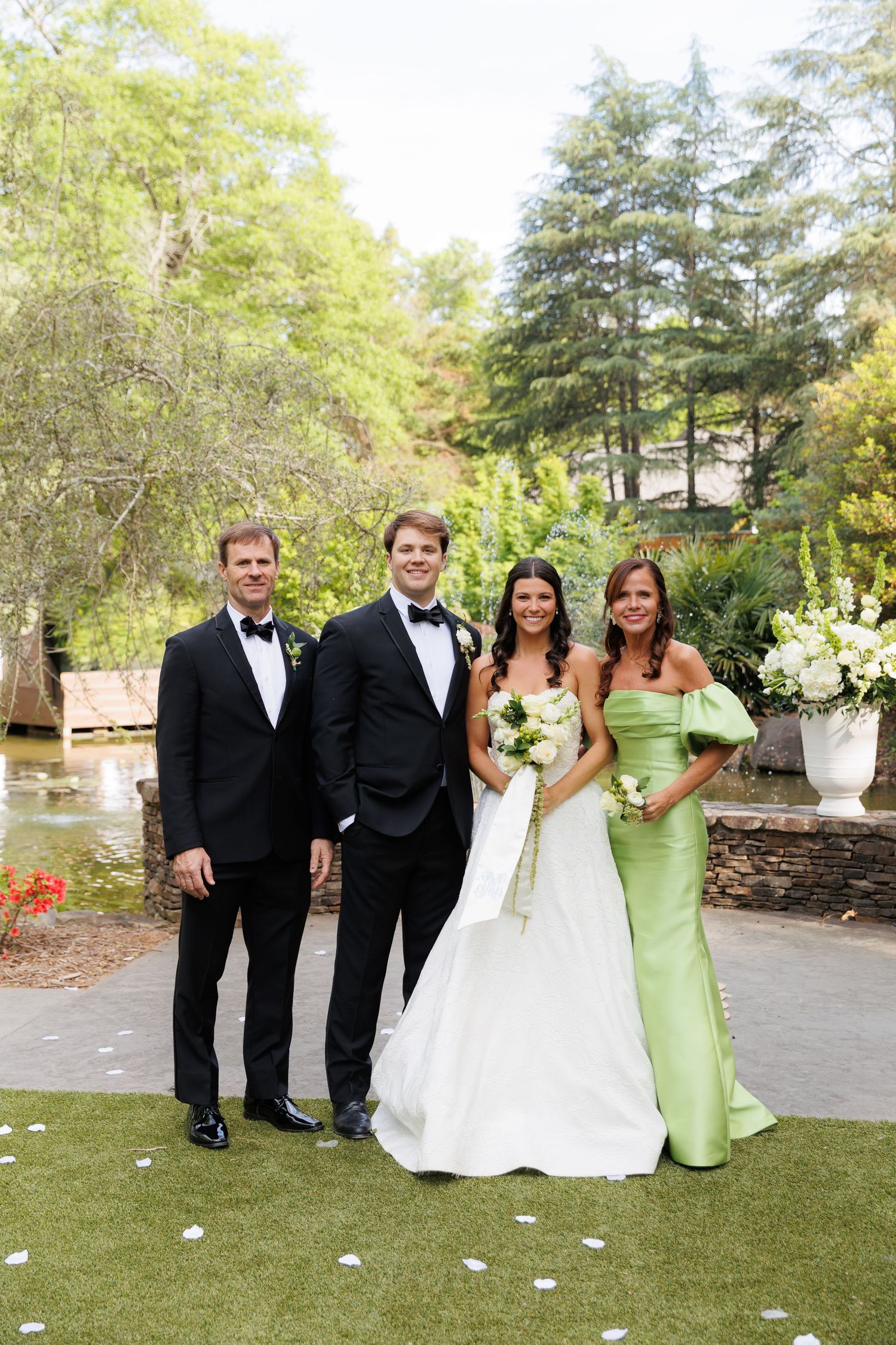 Wedding party of four poses outdoors, bride in white gown, groom in tuxedo, guests in formal attire.