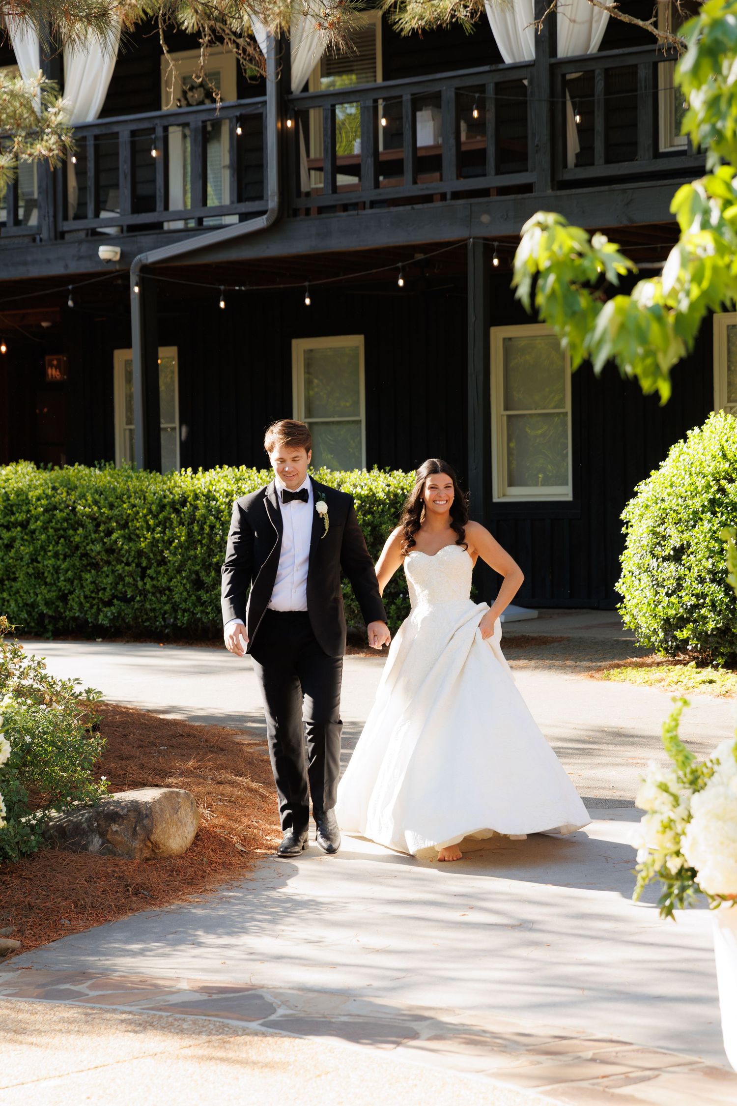 A bride in a white ball gown and groom in a black tuxedo walk hand in hand along a pathway outside an elegant venue.