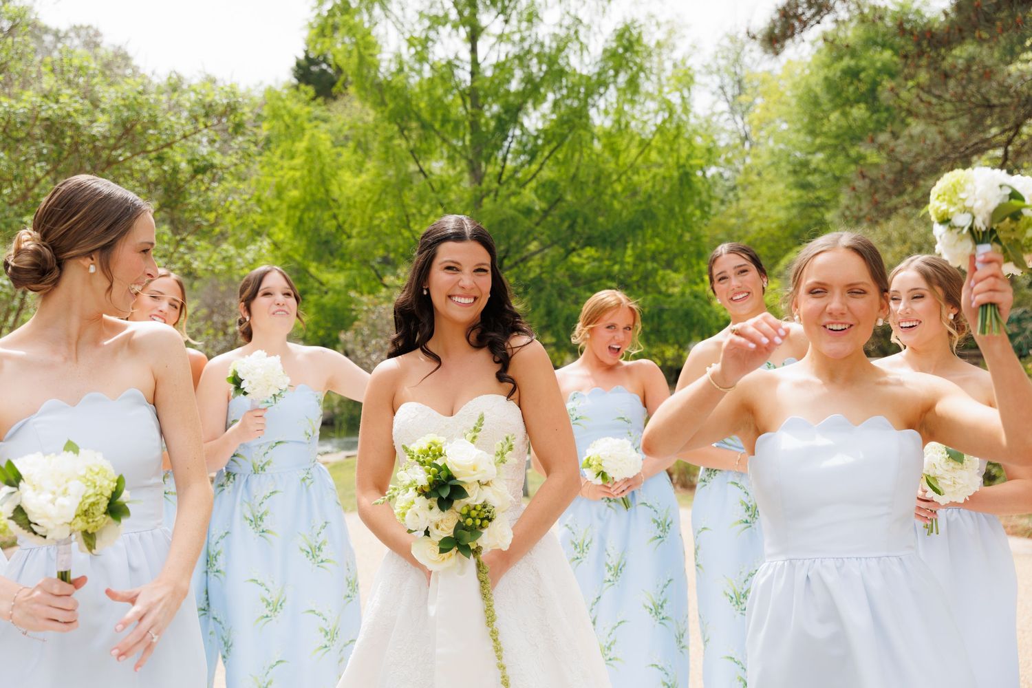Bride in white gown smiles with bridesmaids in light blue floral dresses holding white bouquets outdoors.