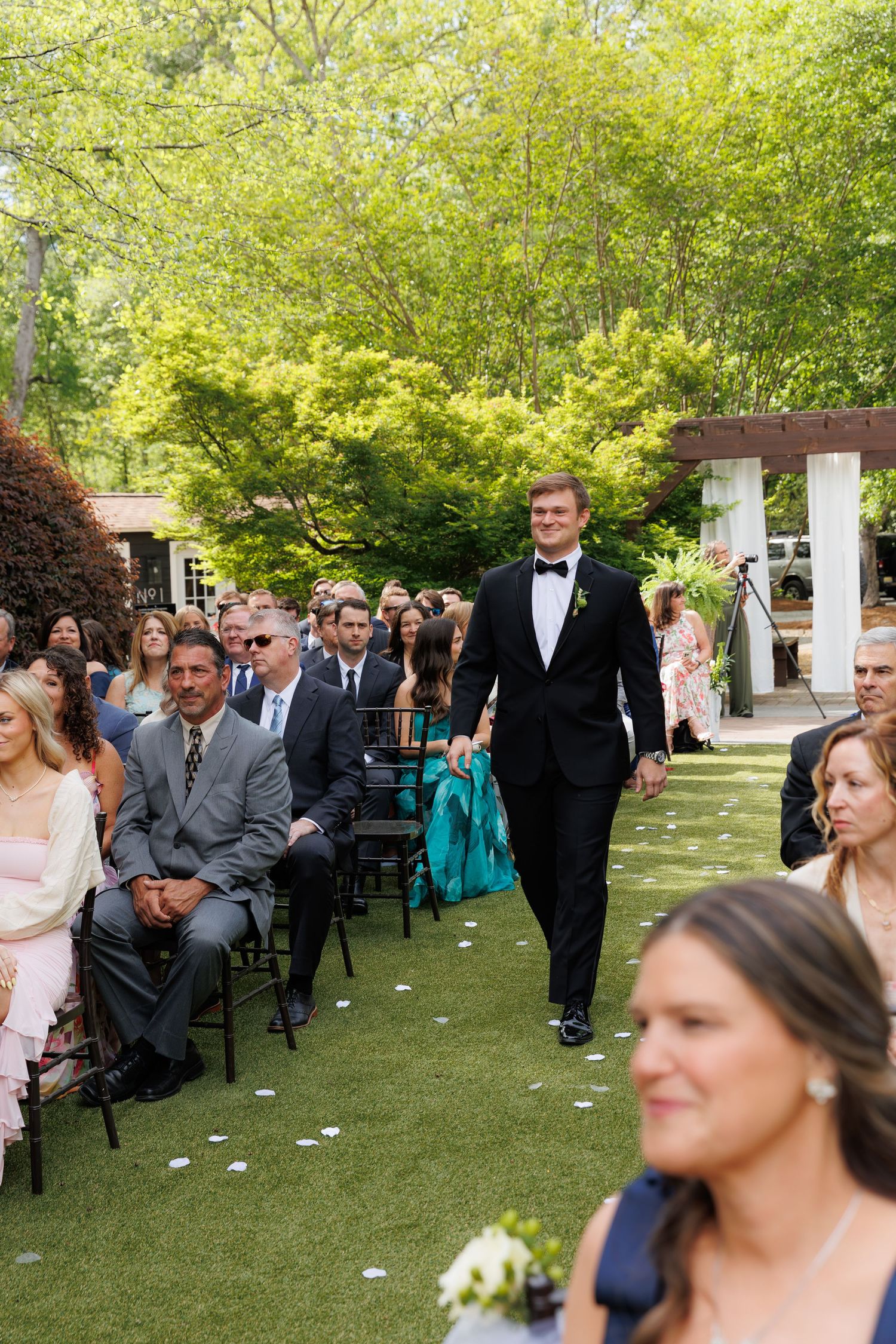 Groom in black tuxedo walks down outdoor wedding aisle surrounded by seated guests in a lush garden setting.