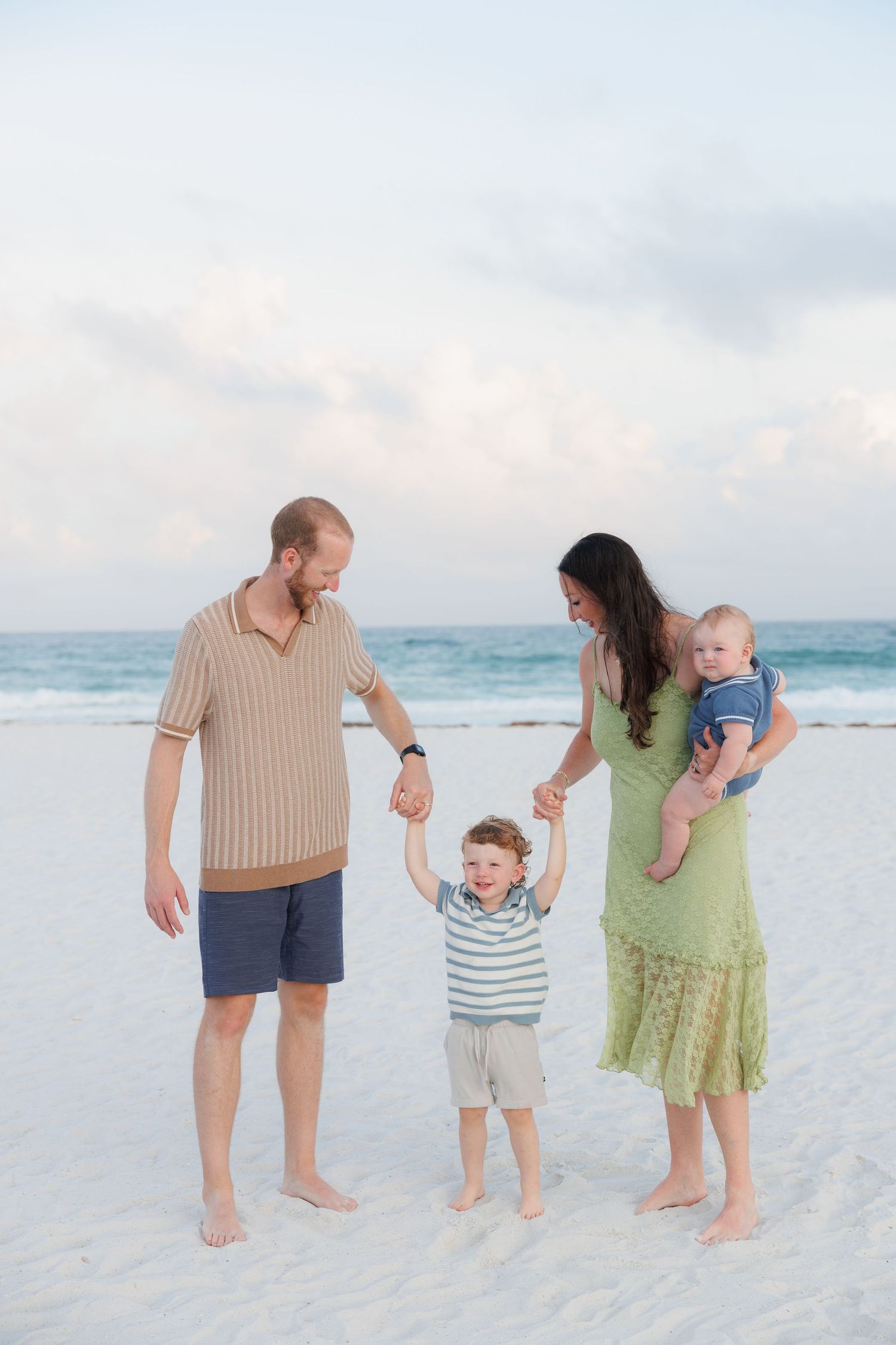 Family of four enjoying a beach day, parents holding toddler's hands while mom carries baby, ocean waves in background.