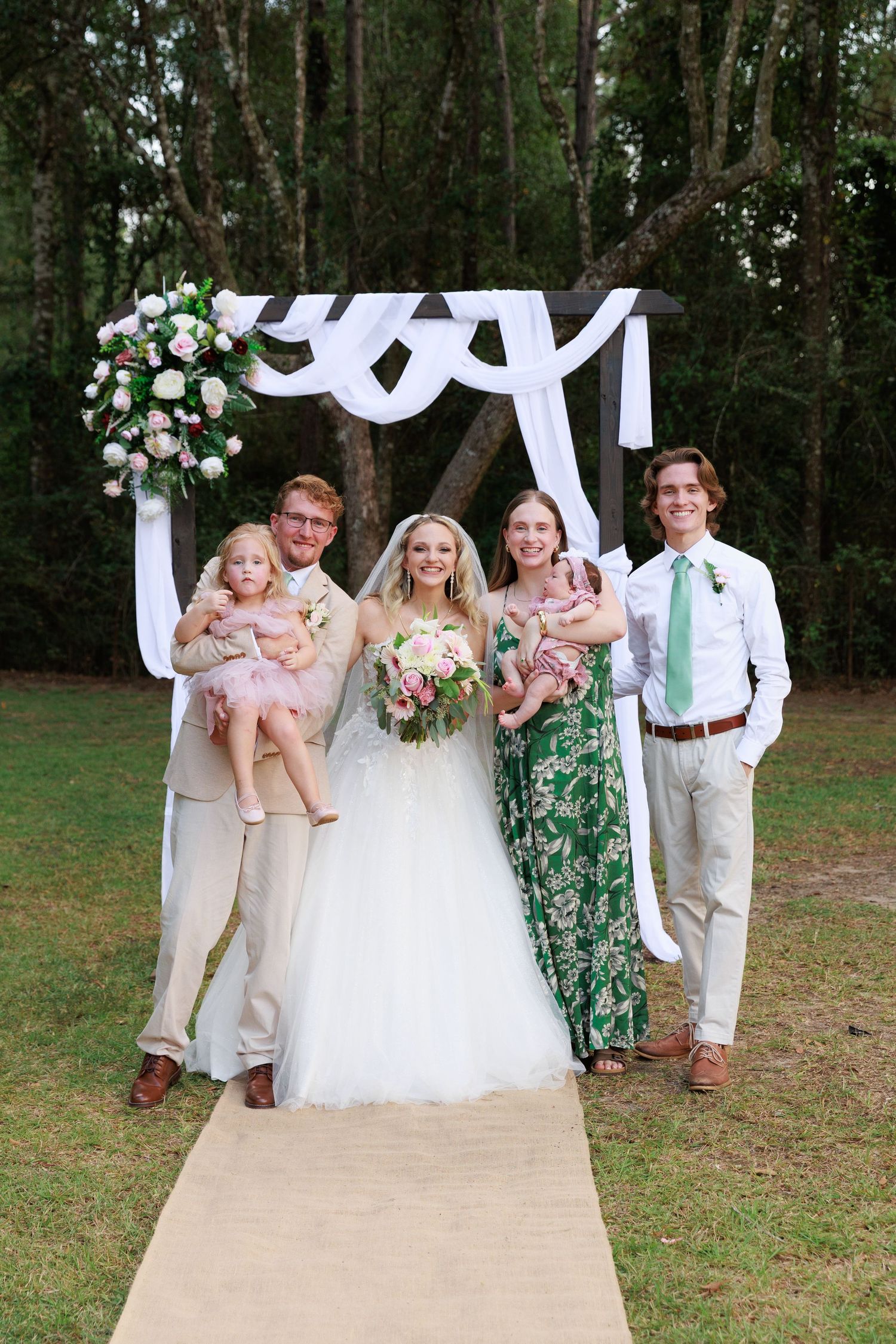 Wedding party poses playfully on outdoor walkway with white decorative arch and trees in background.