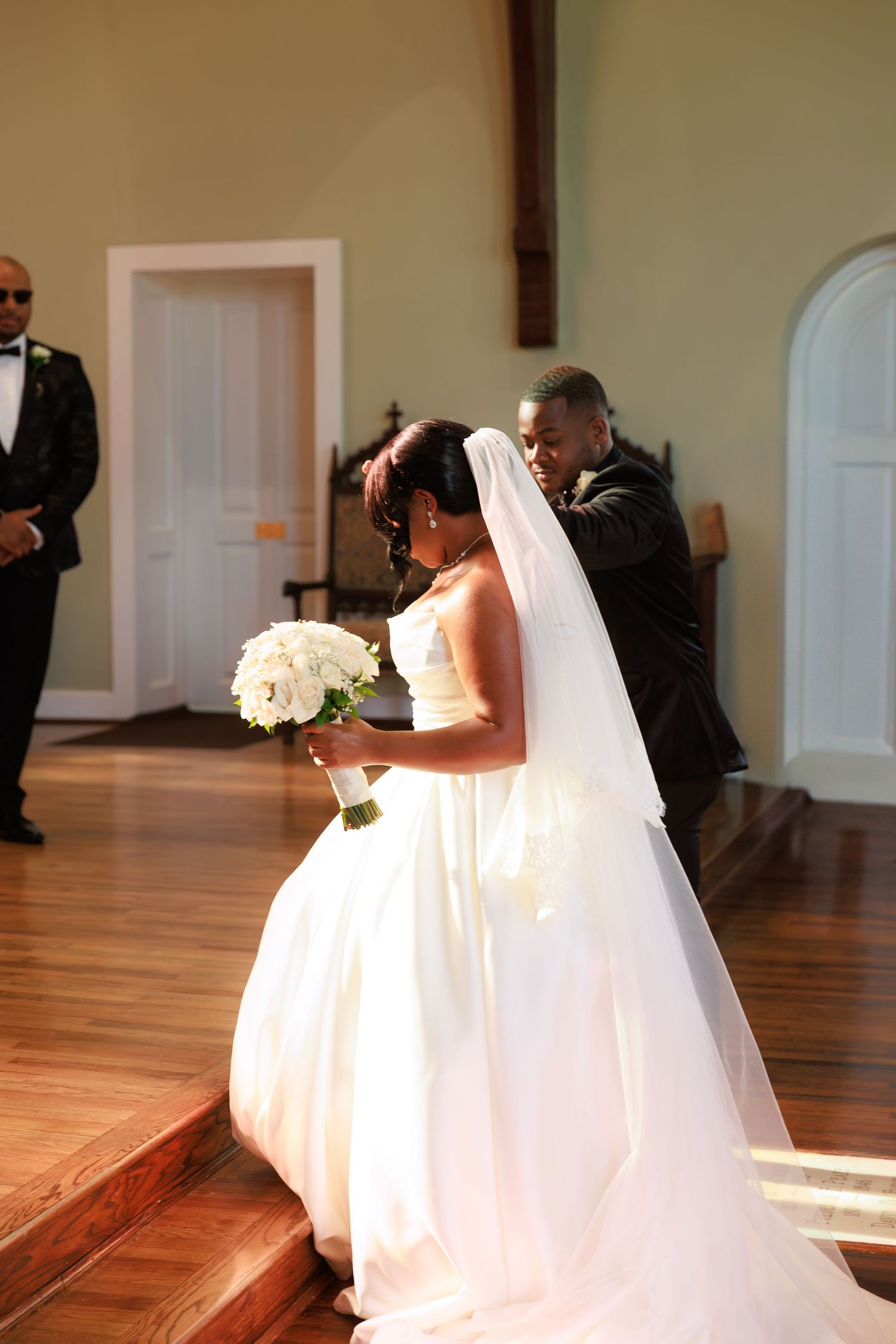 A bride in a white wedding gown with a flowing veil holds a bouquet of white flowers during a church ceremony.