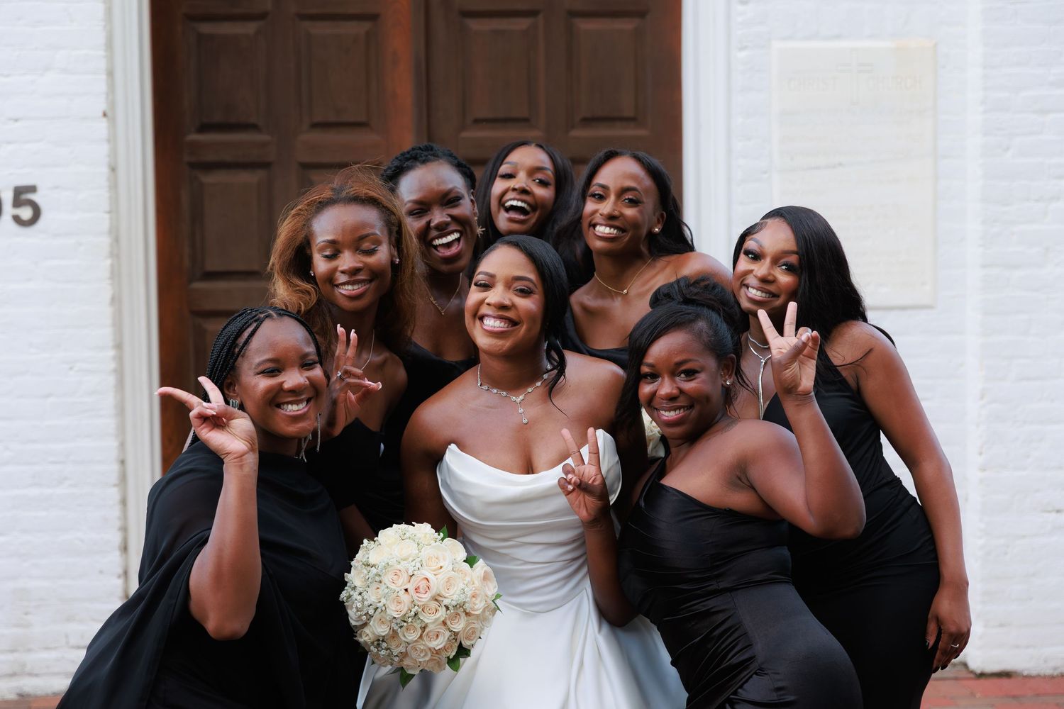 A joyful wedding party in black dresses and one white gown pose together while making peace signs.