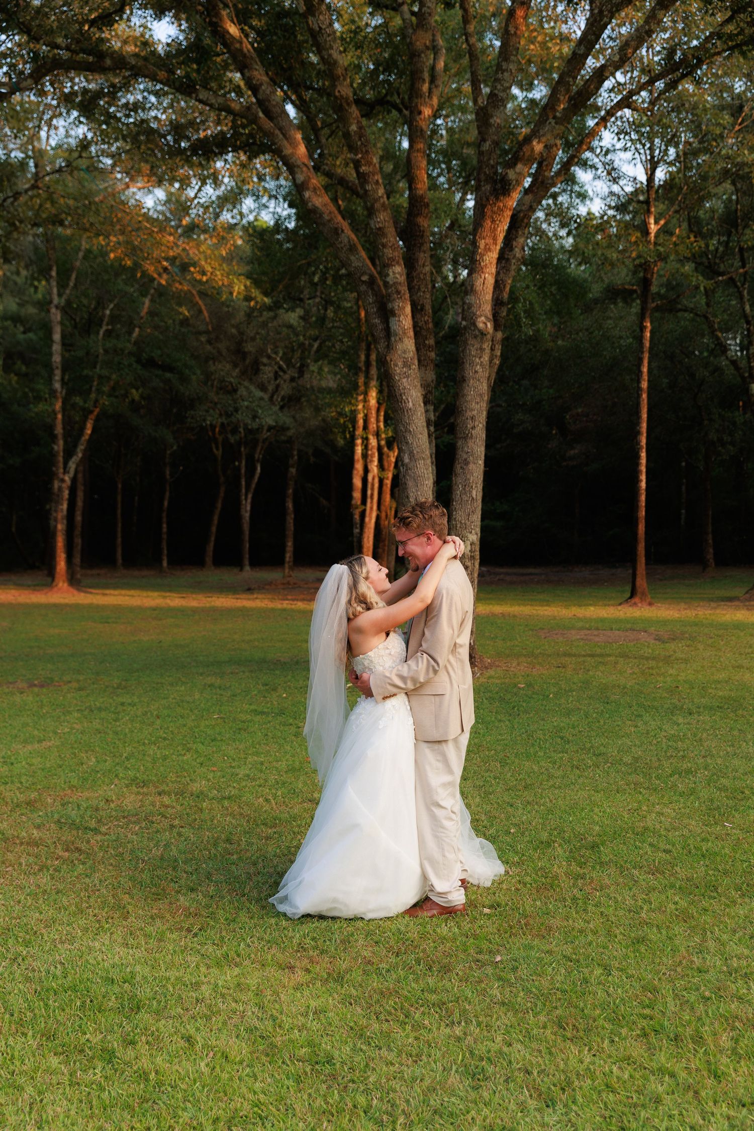 Two people in formal attire dance together in a grassy field at sunset with trees in the background.