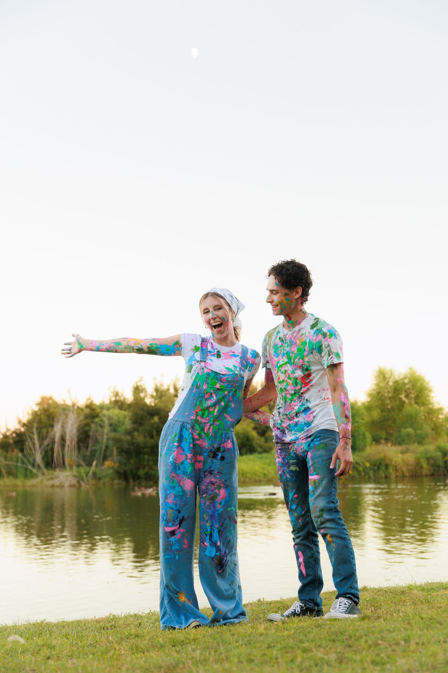 People in paint-splattered denim outfits playing and laughing together by a lake at sunset.