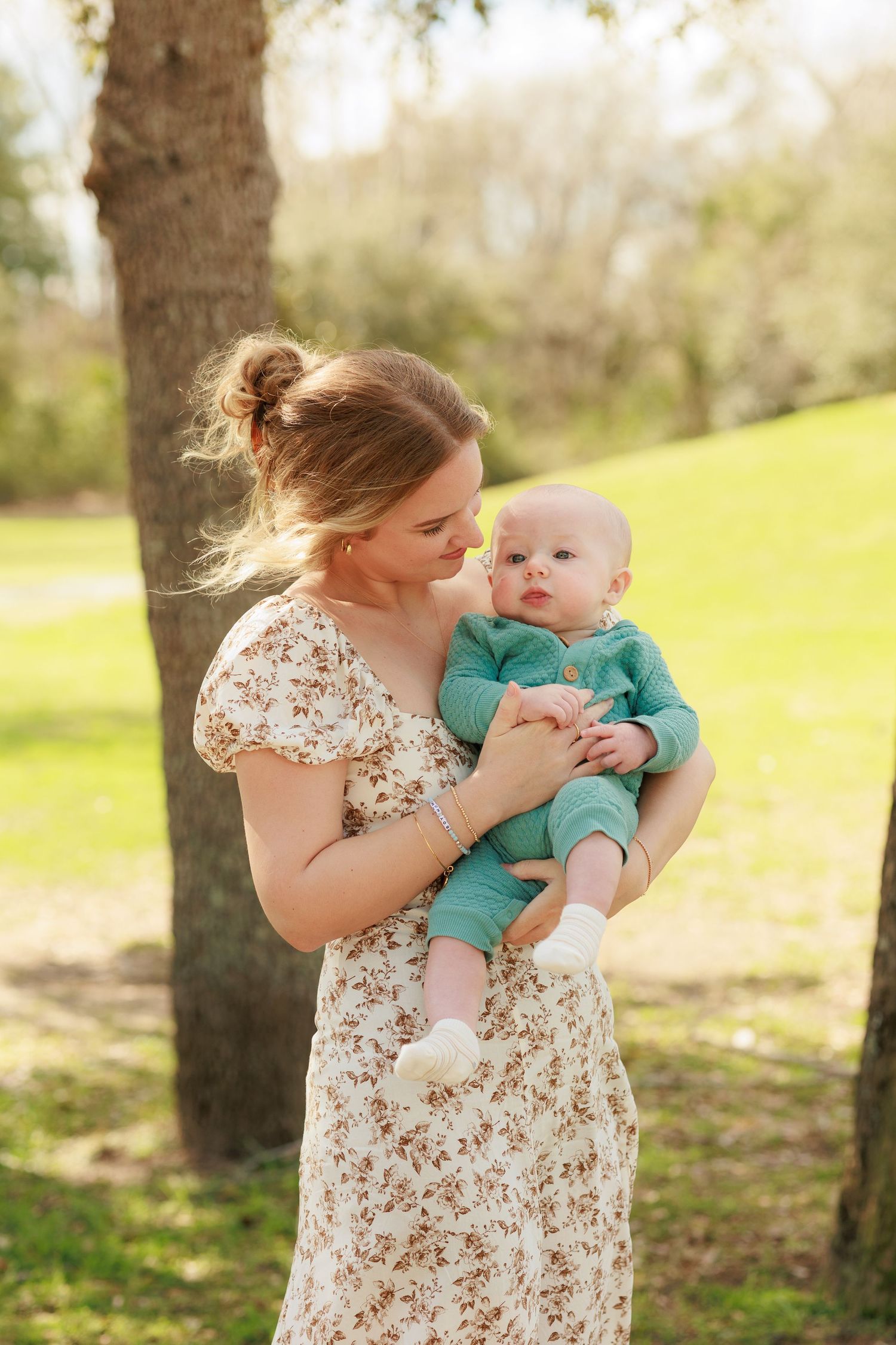 A woman in a floral dress holds a baby wearing teal in a sunny outdoor setting near a tree.