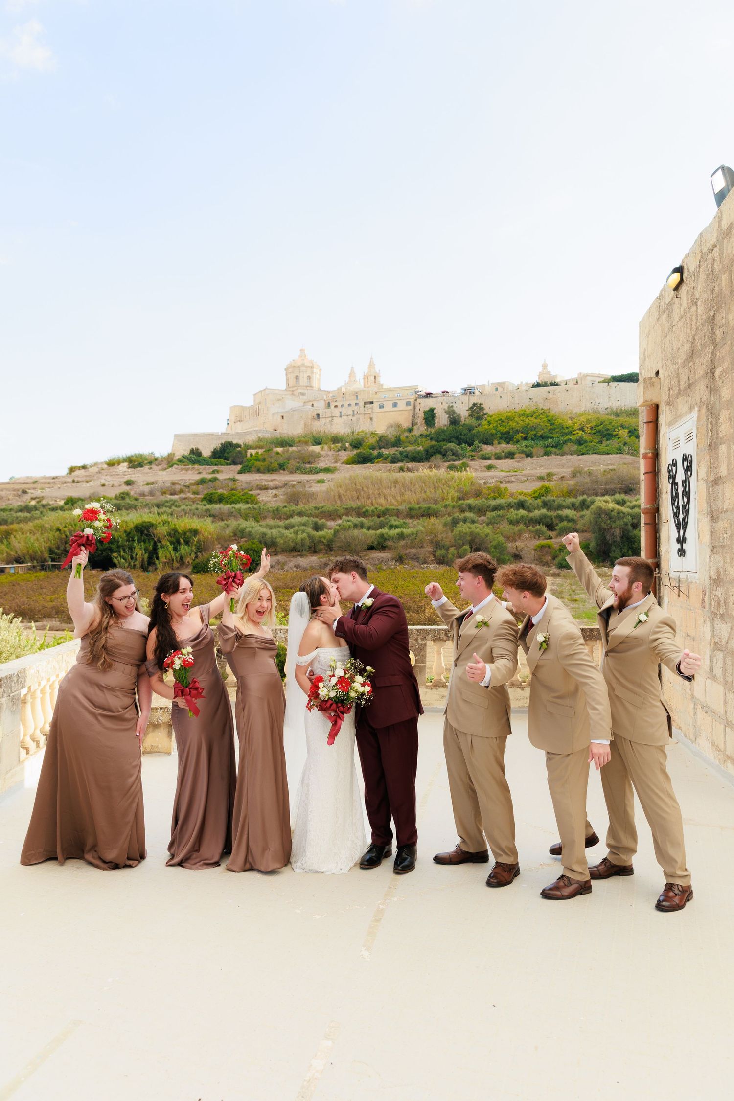 Wedding party in neutral tones poses for creative panoramic photo against white stone wall and scenic backdrop.
