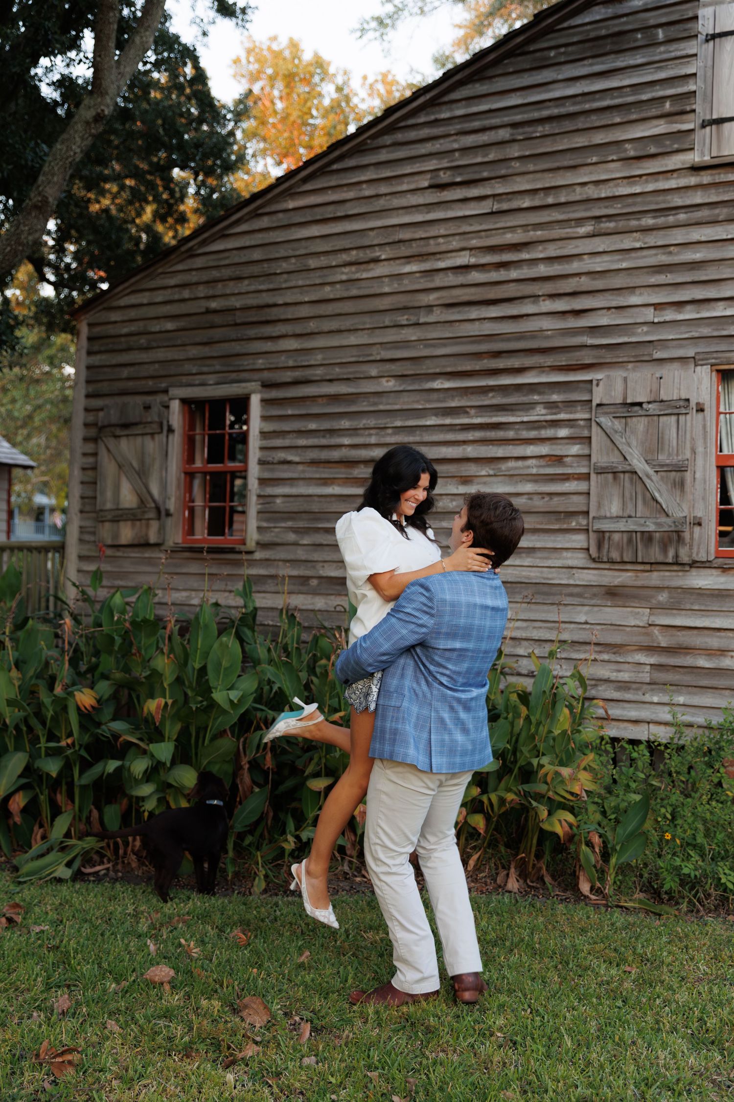 Playful couple dances and spins in front of a rustic wooden barn with autumn foliage in the background.