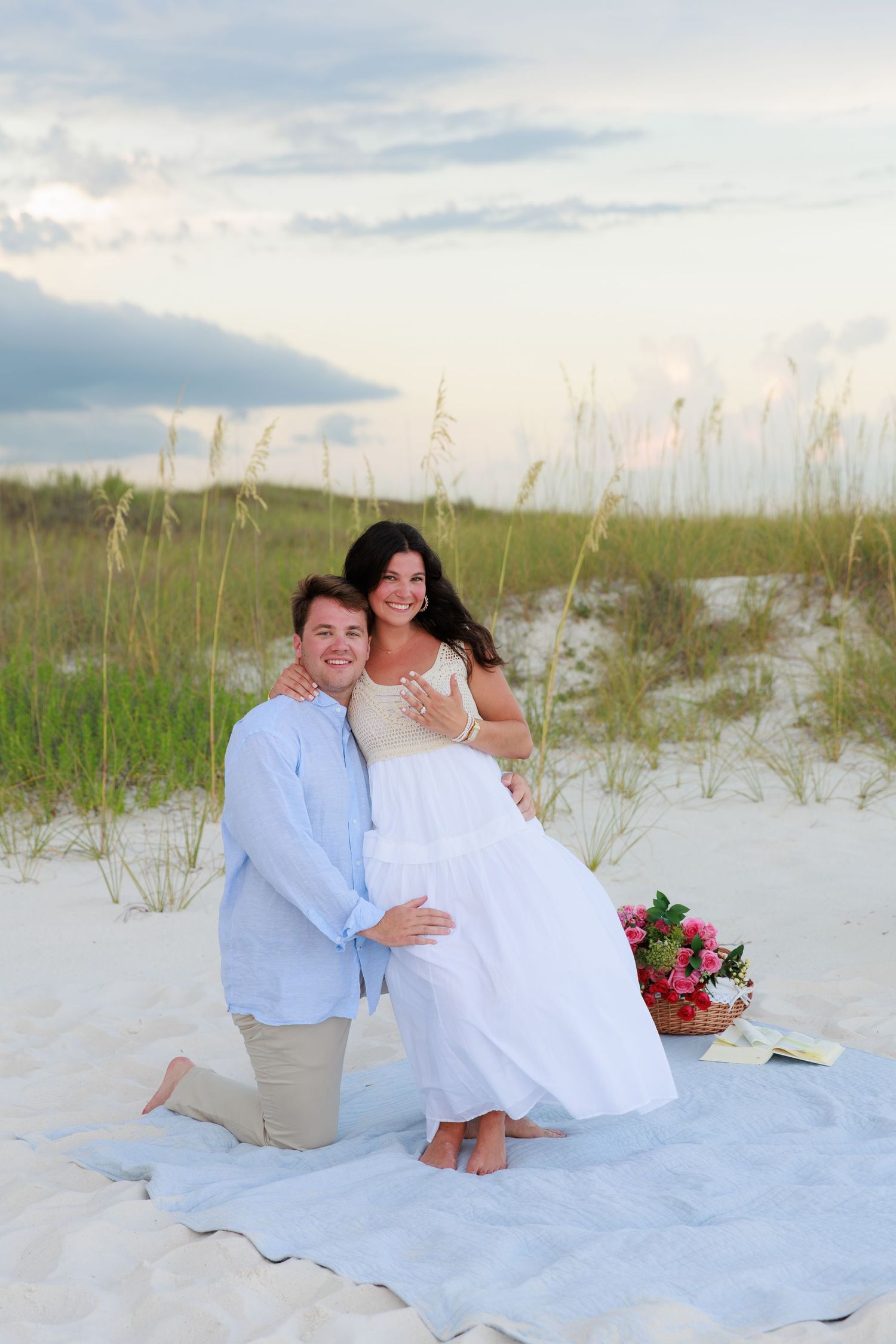A couple shares a romantic moment on a white blanket at the beach with flowers and sea oats in the background.
