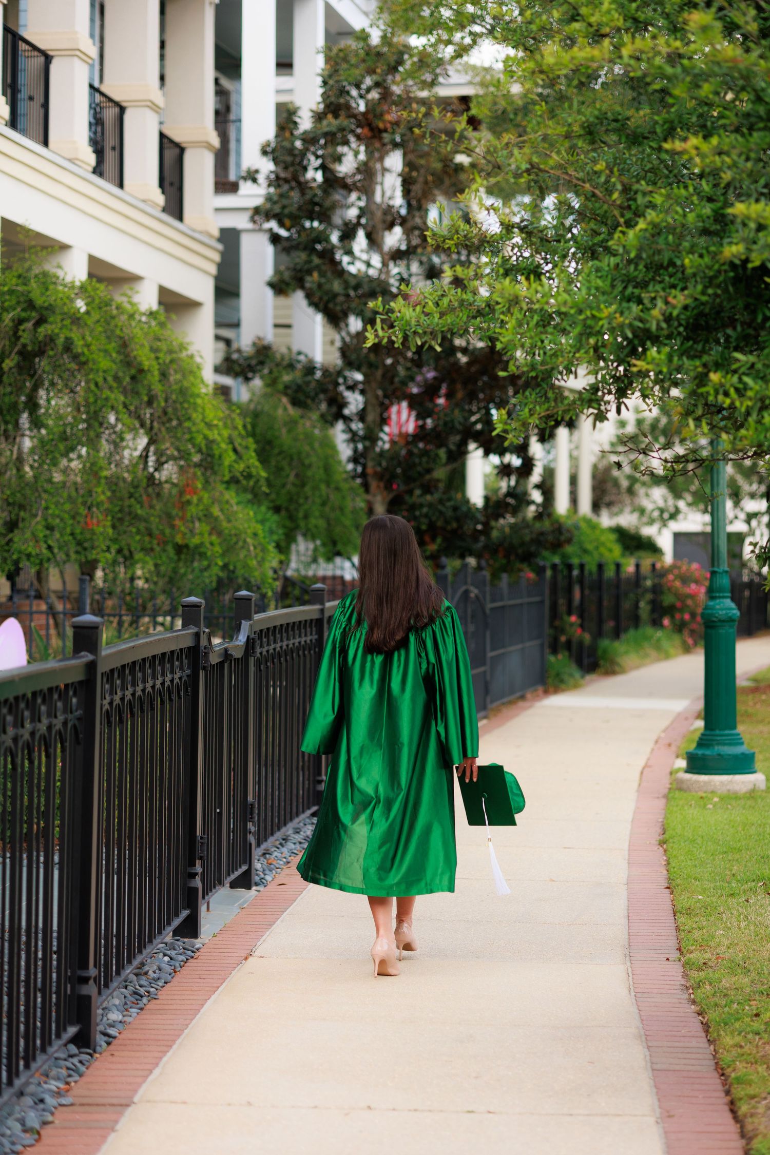 A graduate in a green cap and gown walks along a tree-lined sidewalk beside an elegant white building.