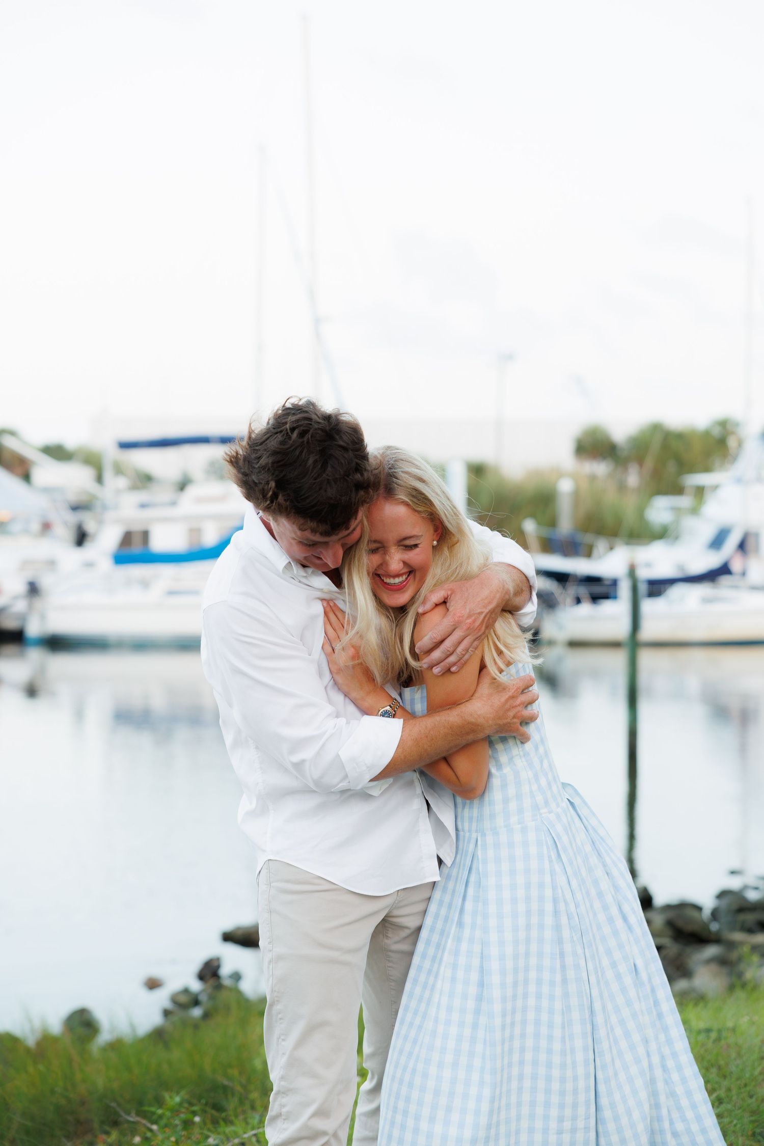 Happy couple hugs and laughs together near a marina with sailboats in background.