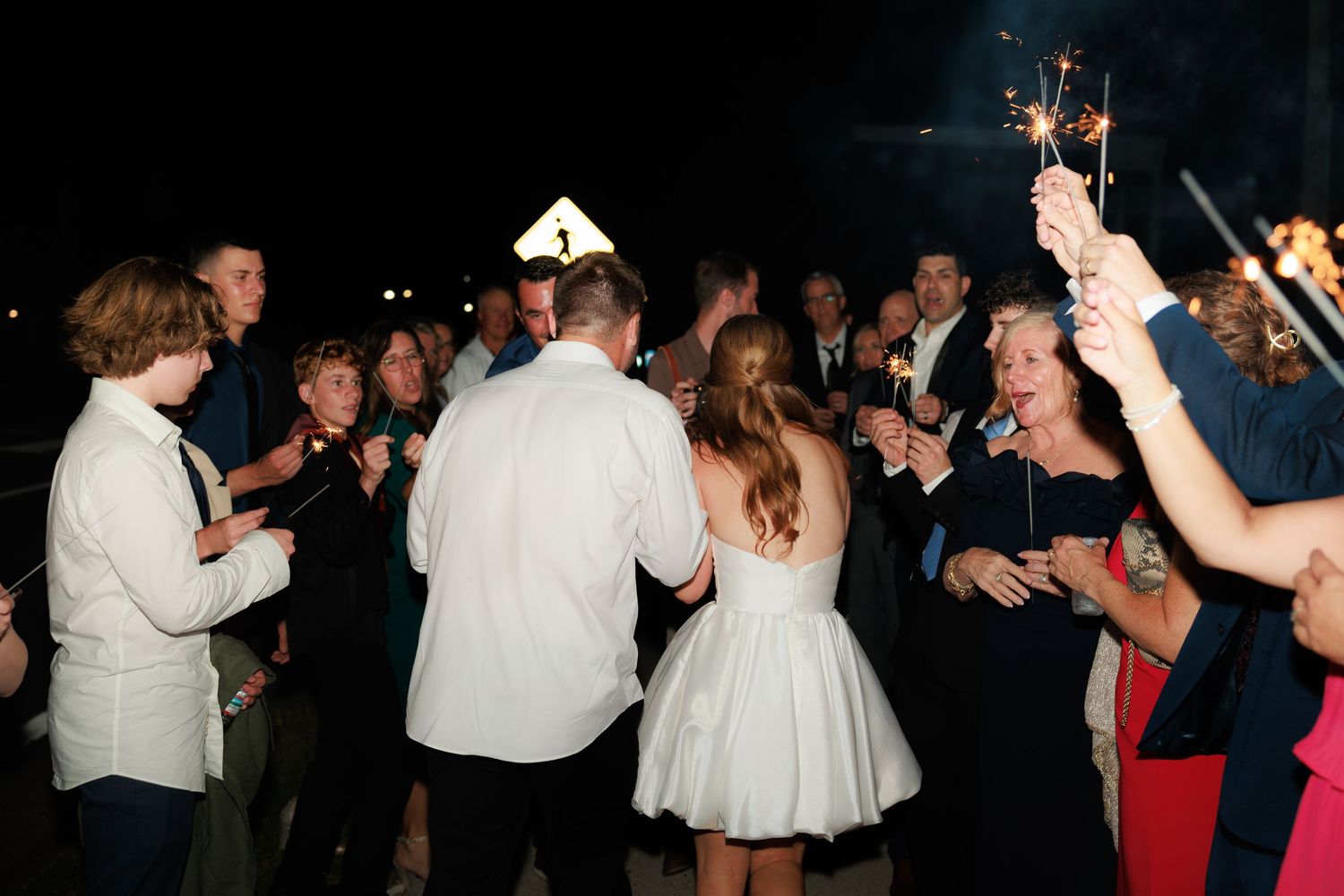 Wedding guests celebrate with sparklers at a nighttime outdoor reception, surrounding the bride and groom in formal attire.