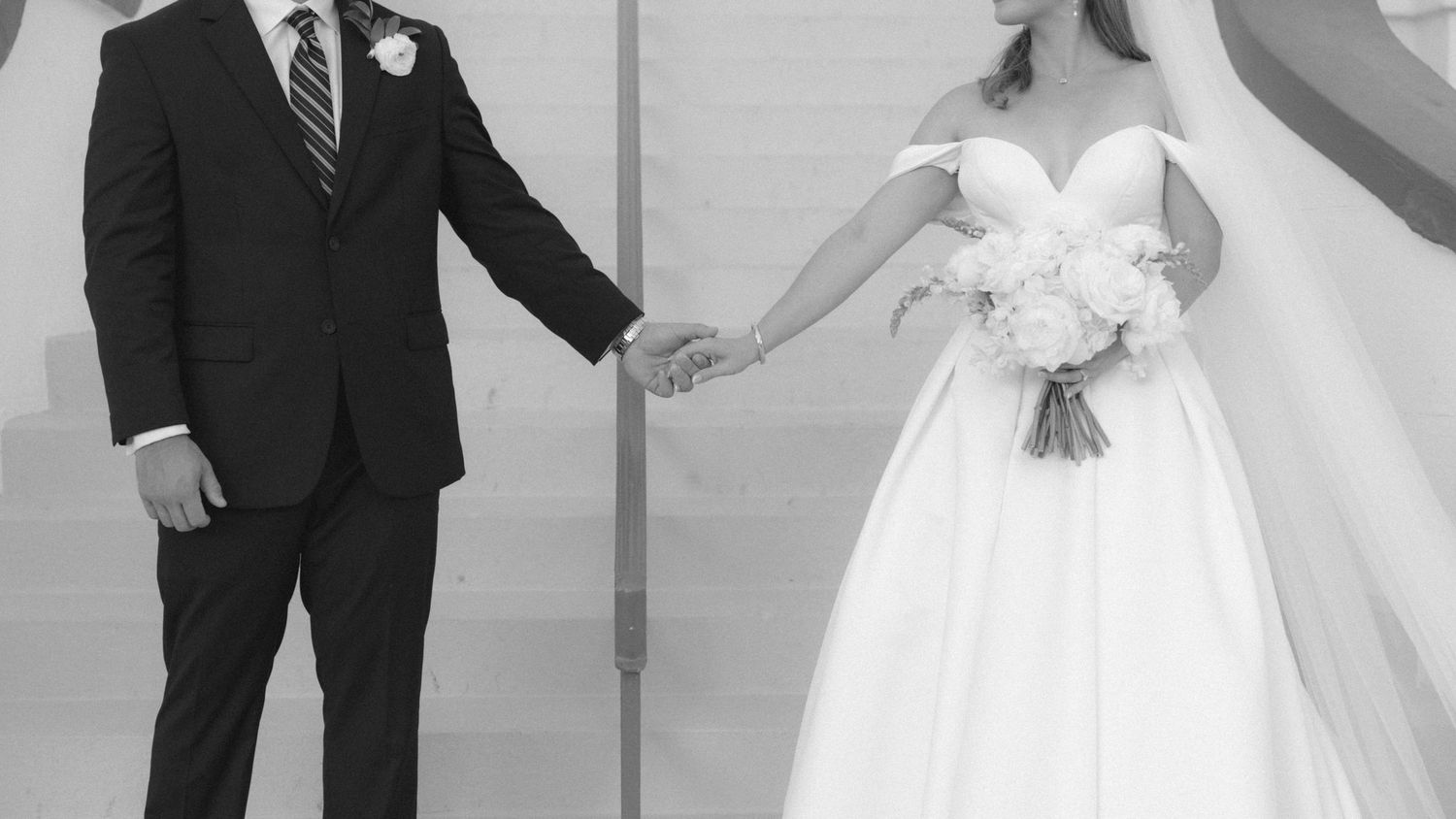Black and white wedding photo of bride in strapless gown holding bouquet and groom in dark suit holding hands.