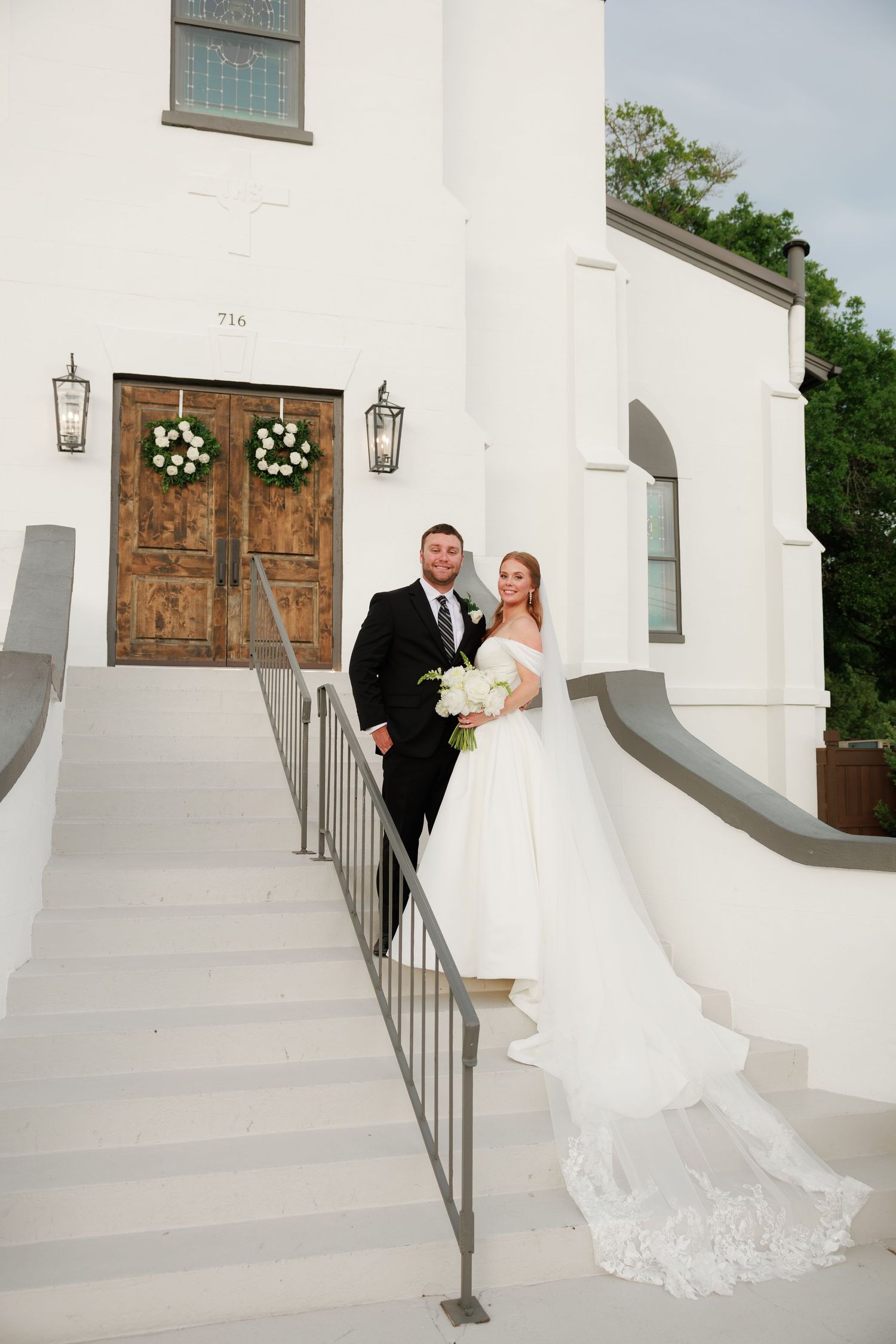 Bride and groom pose on white church steps, bride in elegant gown with lace train holding white bouquet.