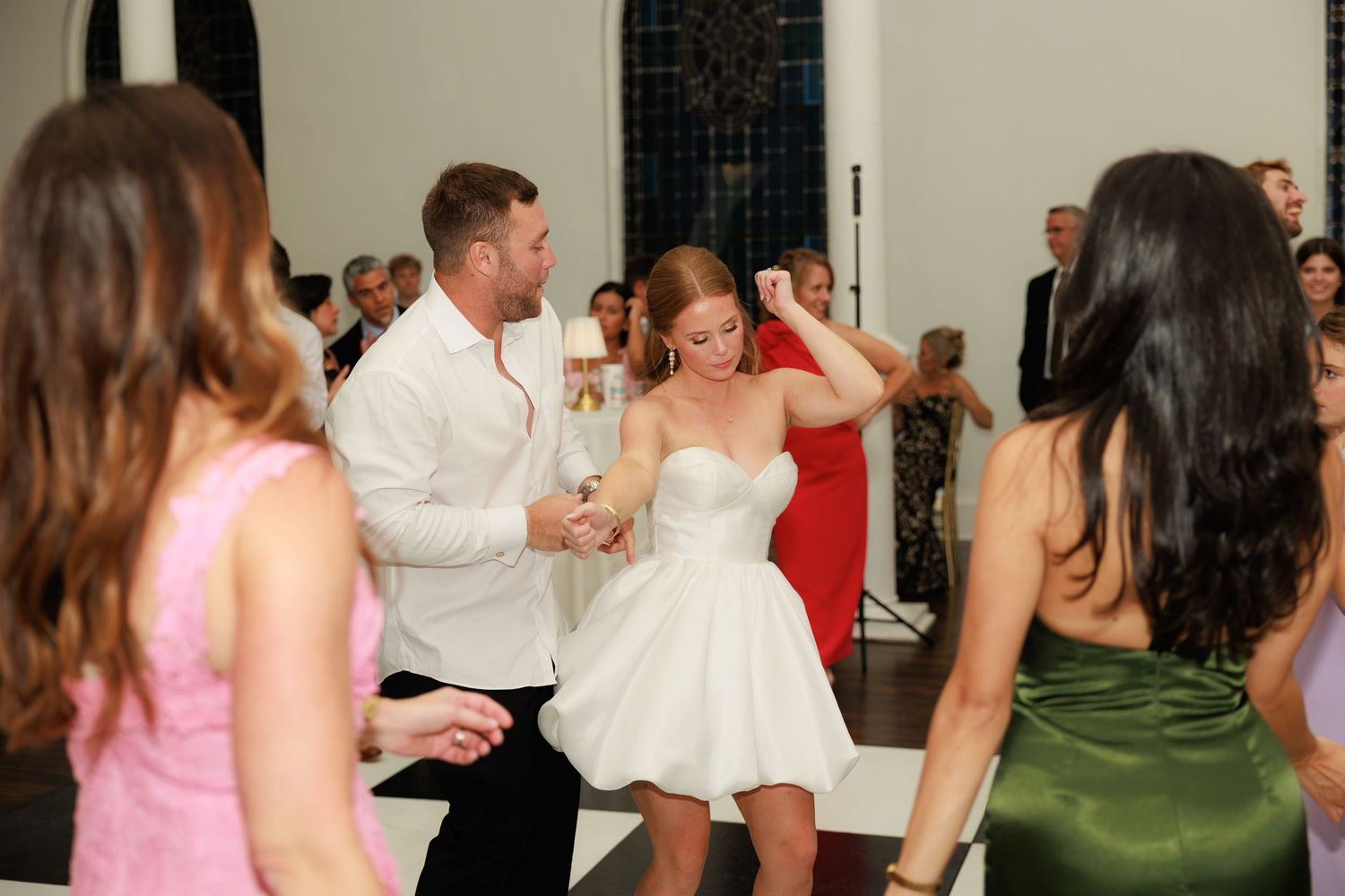 Bride in short white dress dances joyfully with groom at wedding reception surrounded by guests.
