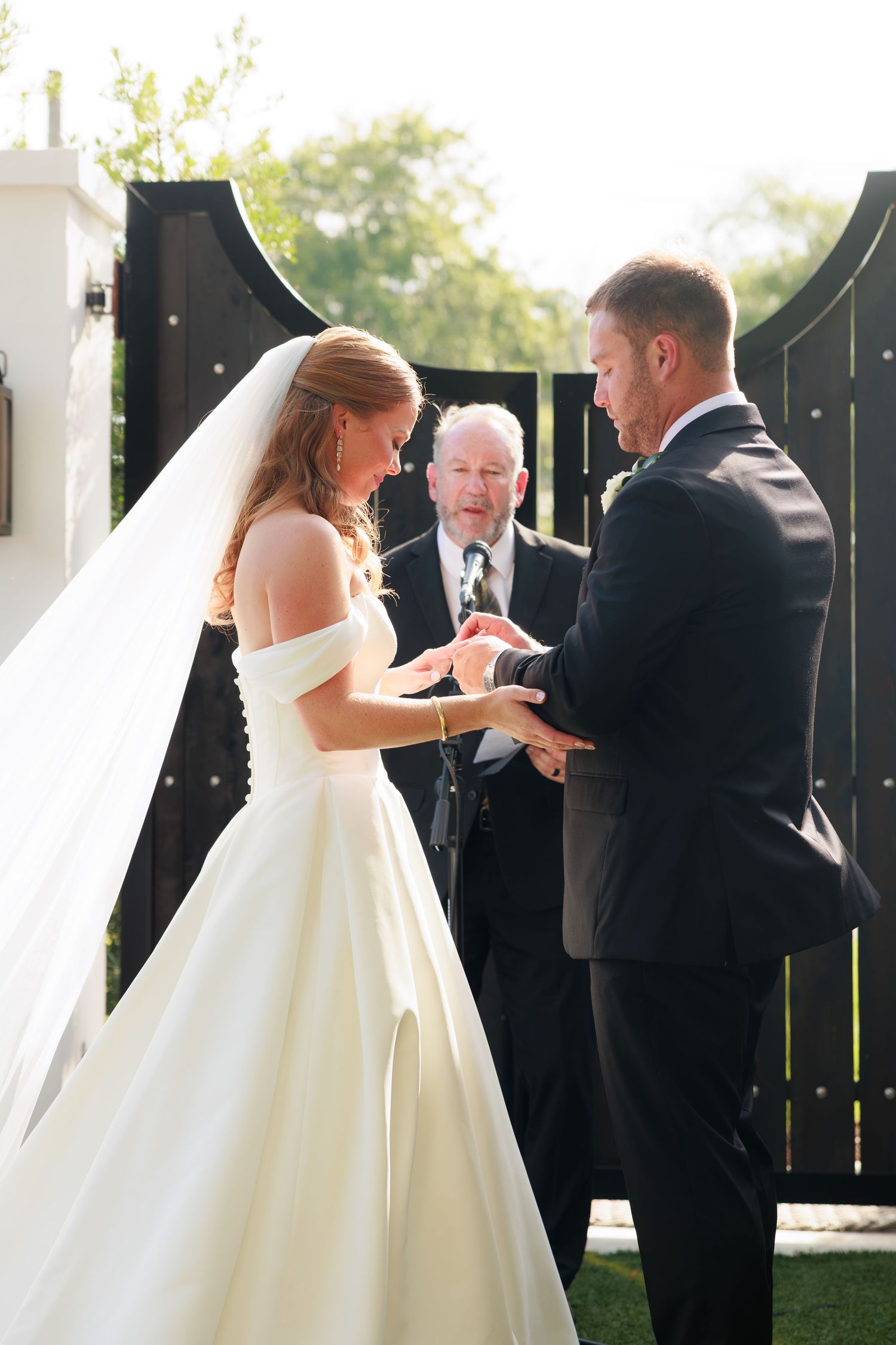 Bride in white gown exchanging rings with groom during outdoor wedding ceremony with officiant.