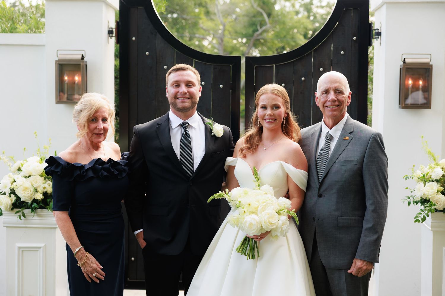 Wedding party of four poses before elegant black gates with floral arrangements, bride holding white bouquet in strapless gown.
