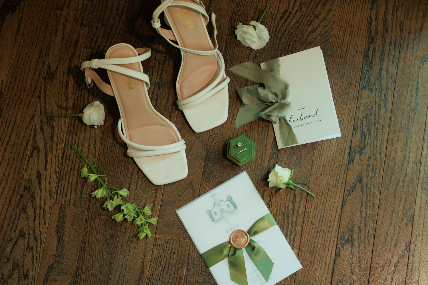 White strappy bridal heels beside green-ribboned stationery, floral boutonniere, and eucalyptus on wood floor.