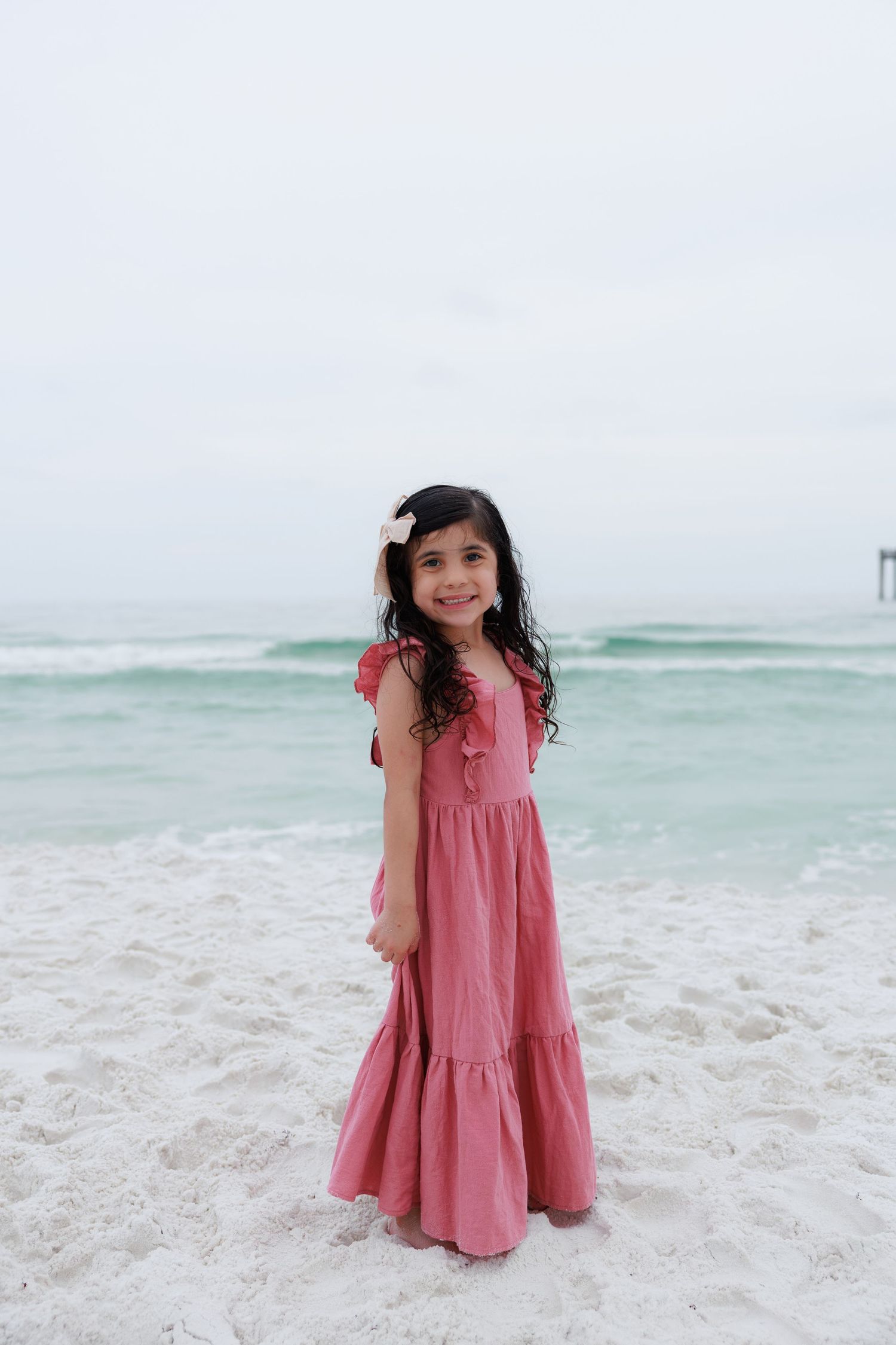 Young girl in pink maxi dress with flower hair clip smiling on white sand beach with ocean behind her.