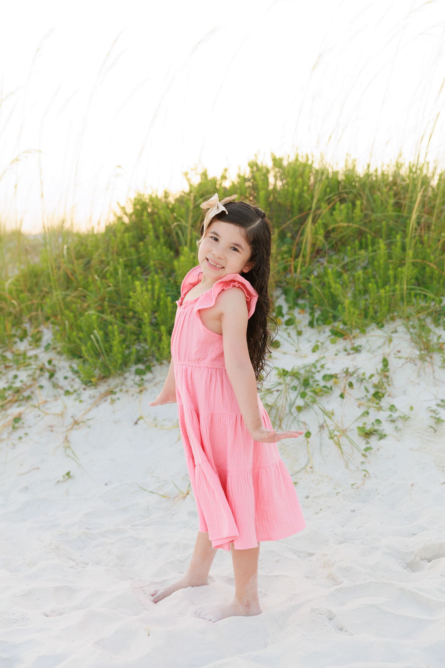 Young girl in pink dress standing barefoot on white sand beach dunes with green sea oats in background.