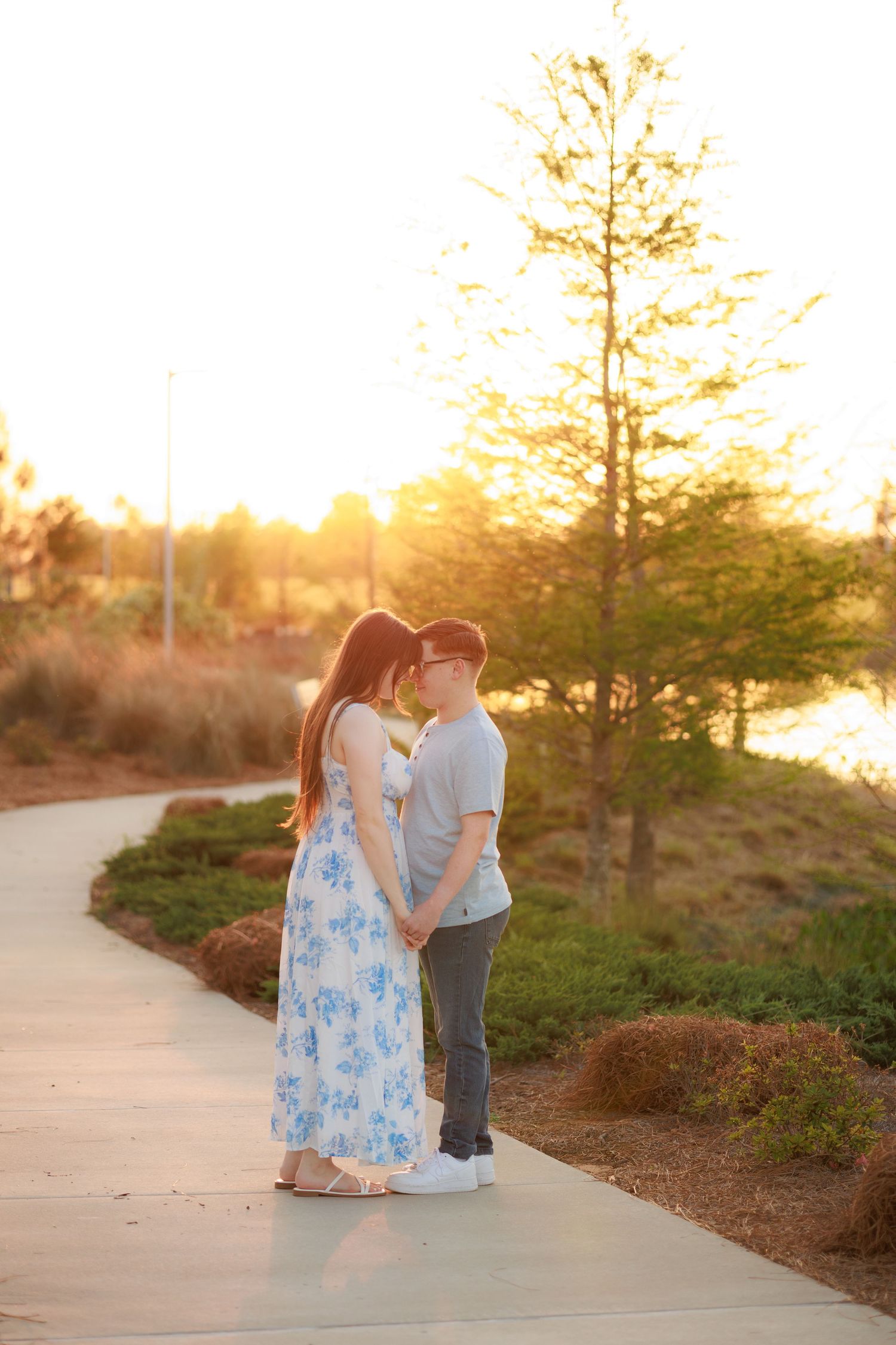 Couple sharing a romantic moment on a sunlit park path at golden hour, woman in floral maxi dress holding hands.