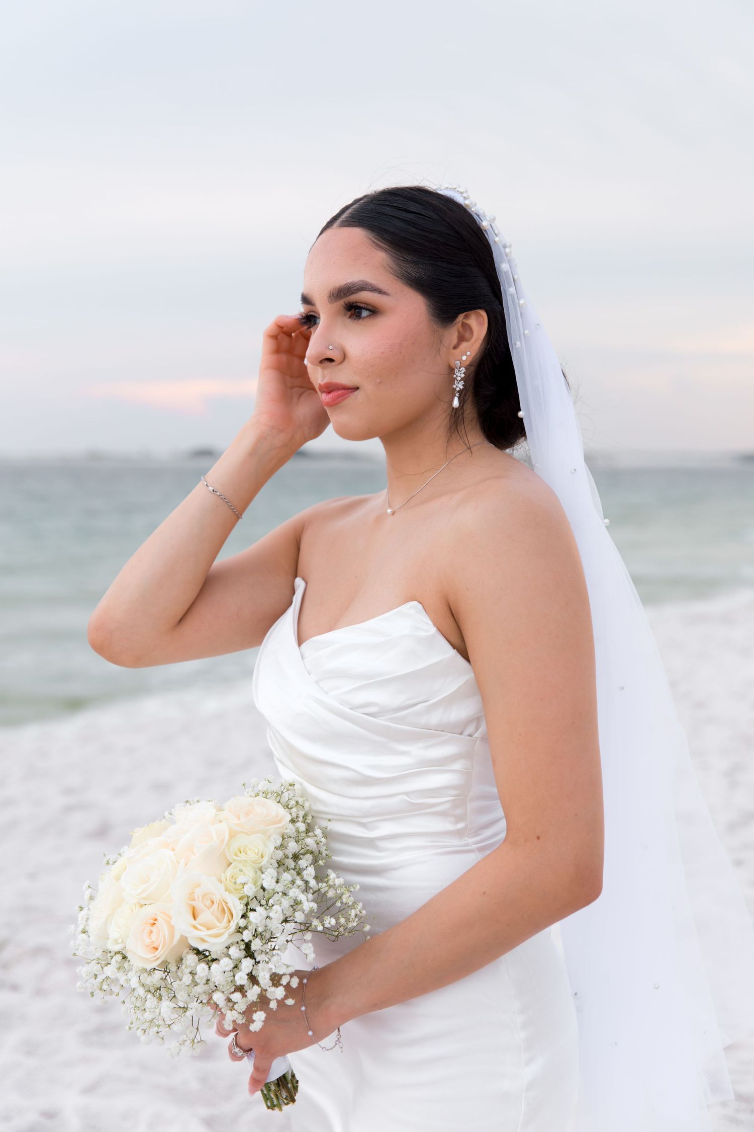Bride in strapless white wedding dress holding bouquet on beach during sunset.