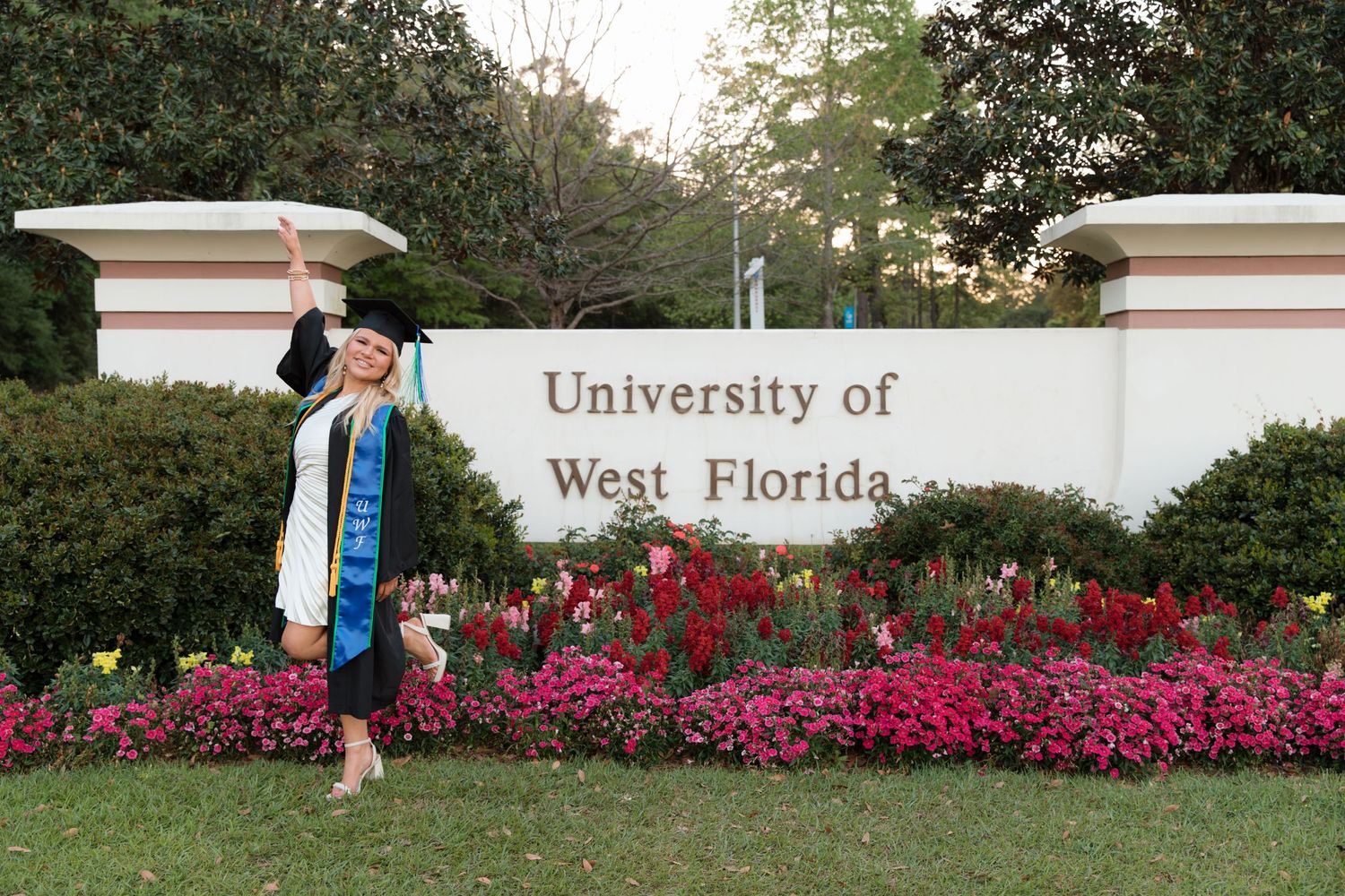 Graduate celebrating at University of West Florida entrance sign with pink flowers.