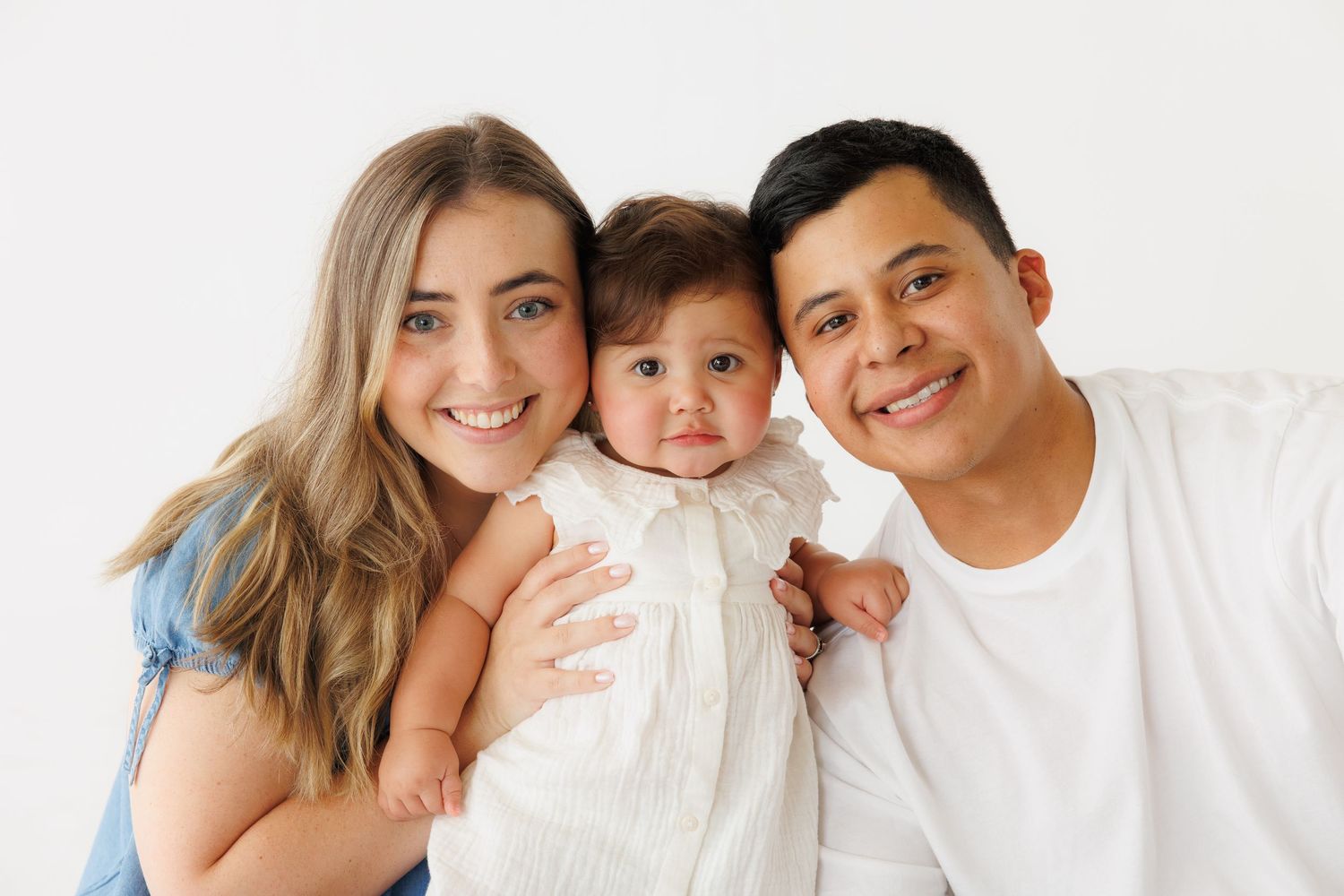 A smiling young family with a mother, father, and baby dressed in white posing together against a white background.
