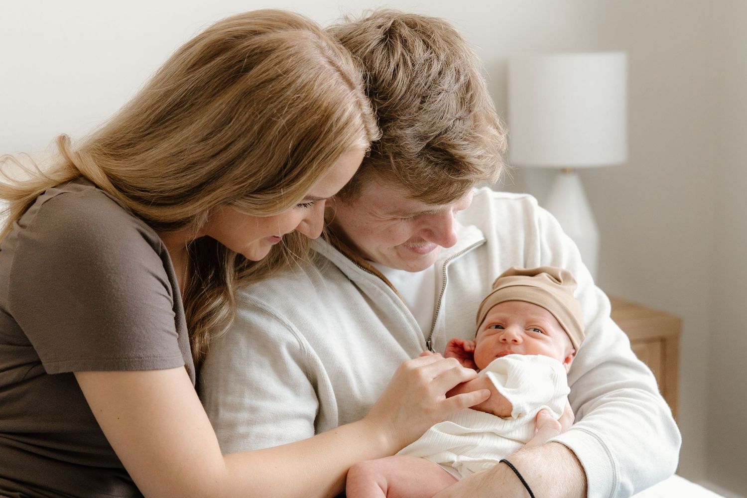 Parents lovingly embrace their newborn baby while sitting together on a couch in a bright, sunlit room.