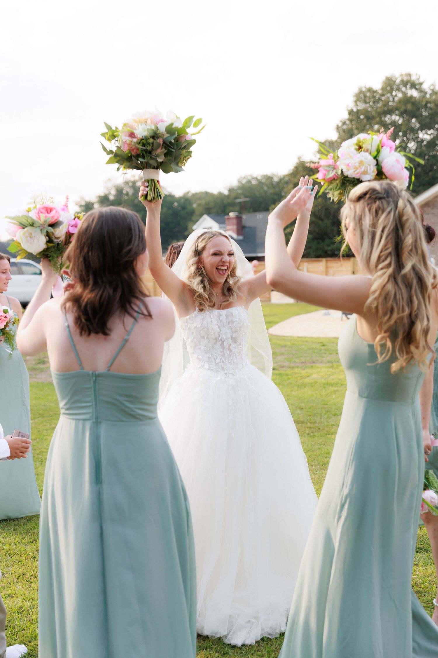 Bridesmaids in sage green dresses celebrate joyfully with bouquets on a sunny outdoor lawn.