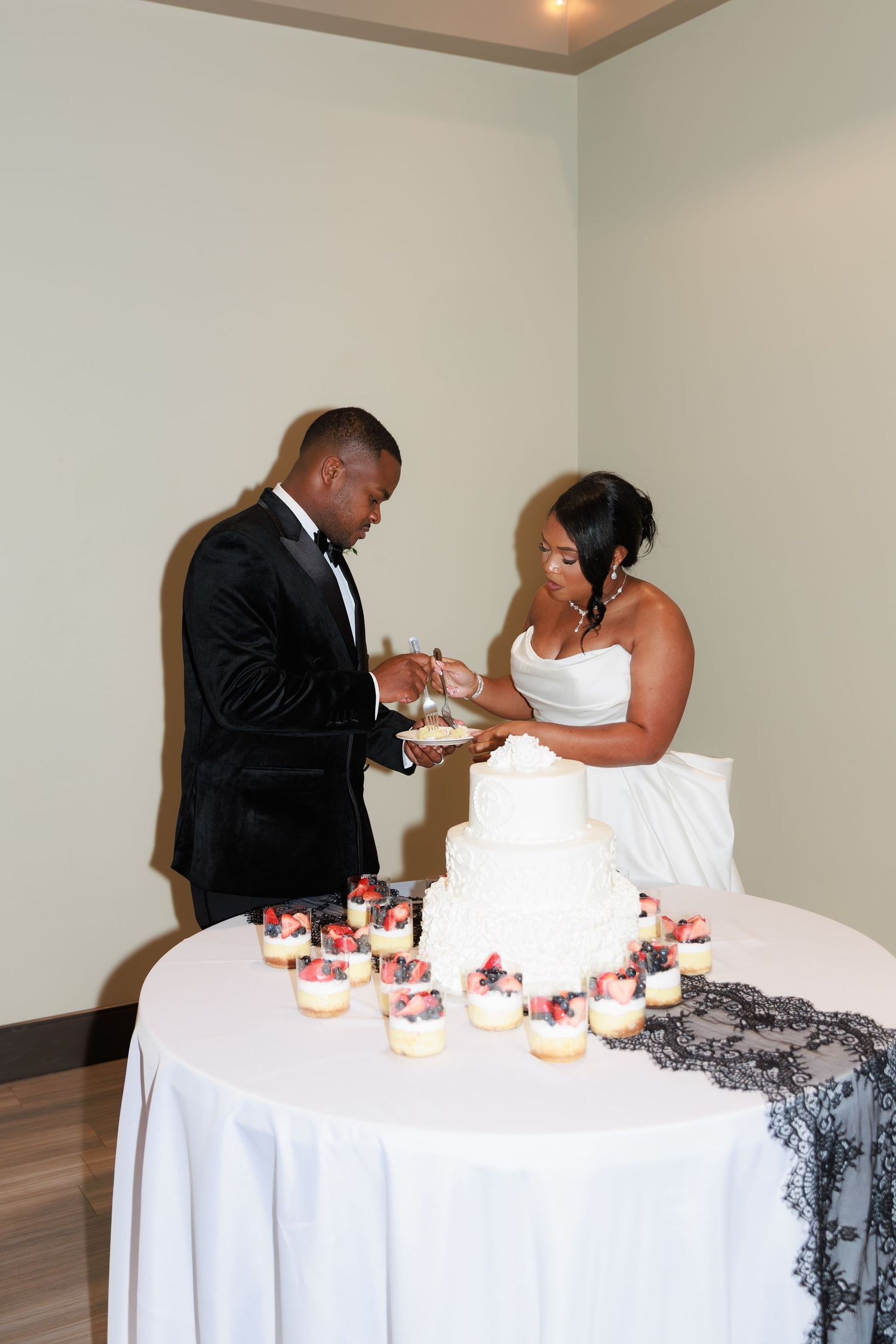 Newlyweds cutting their white wedding cake together with cupcakes displayed on the dessert table.