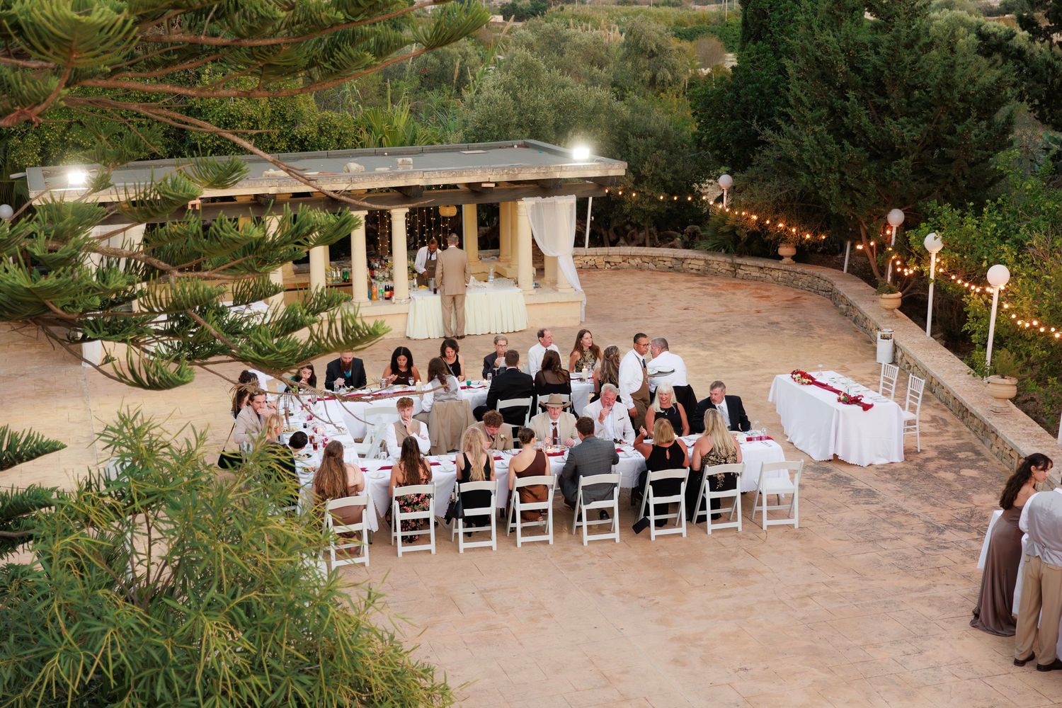 Wedding ceremony setup with white chairs on a stone patio surrounded by lush gardens and string lights overhead.