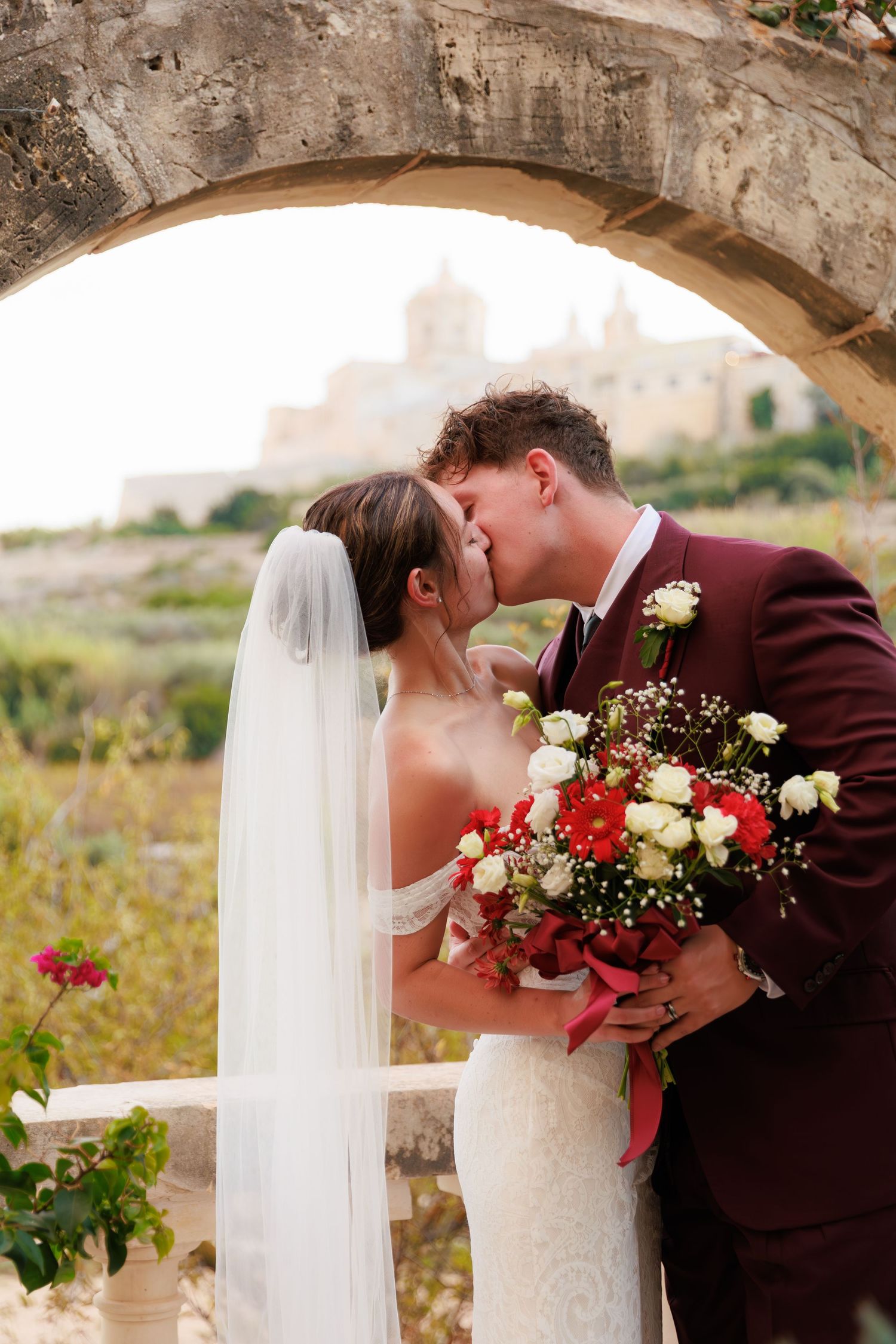 A romantic wedding couple embraces under stone archways with a burgundy suit and white dress against a natural backdrop.