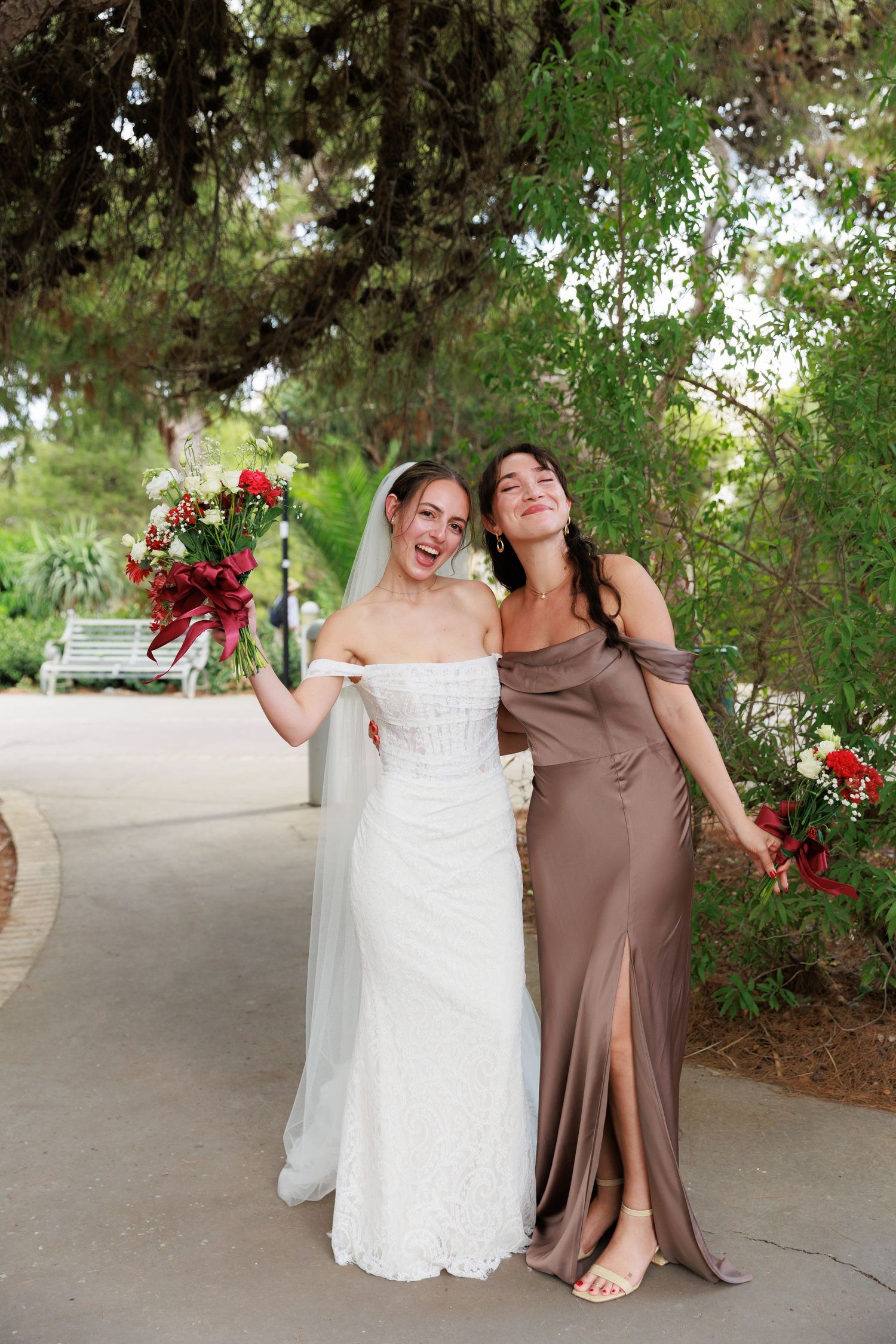 Two wedding attendees share a candid laughing moment on a garden path while holding red floral bouquets.