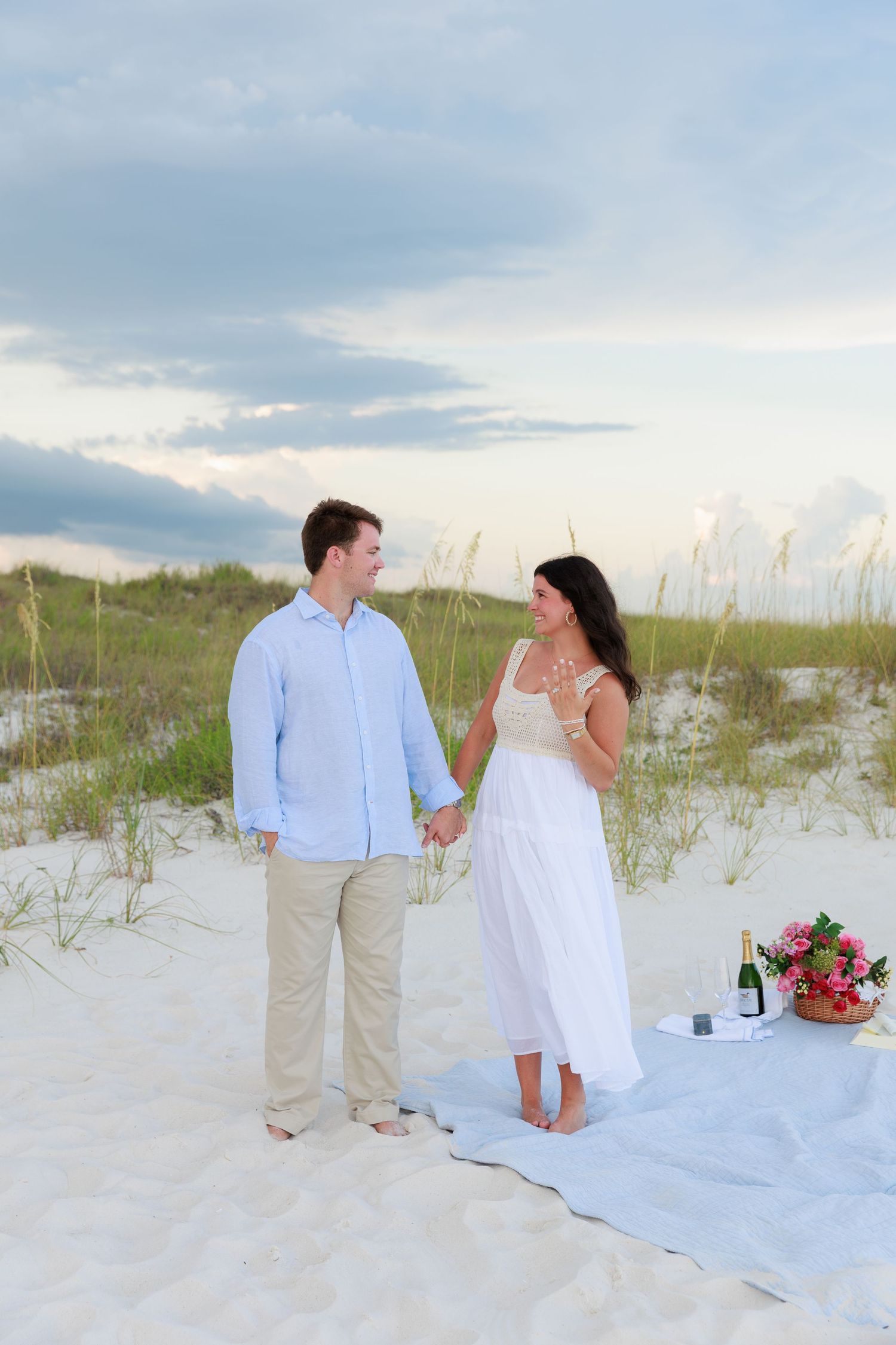 A couple enjoys a romantic beach picnic on white sand dunes at sunset with pink flowers and blanket.