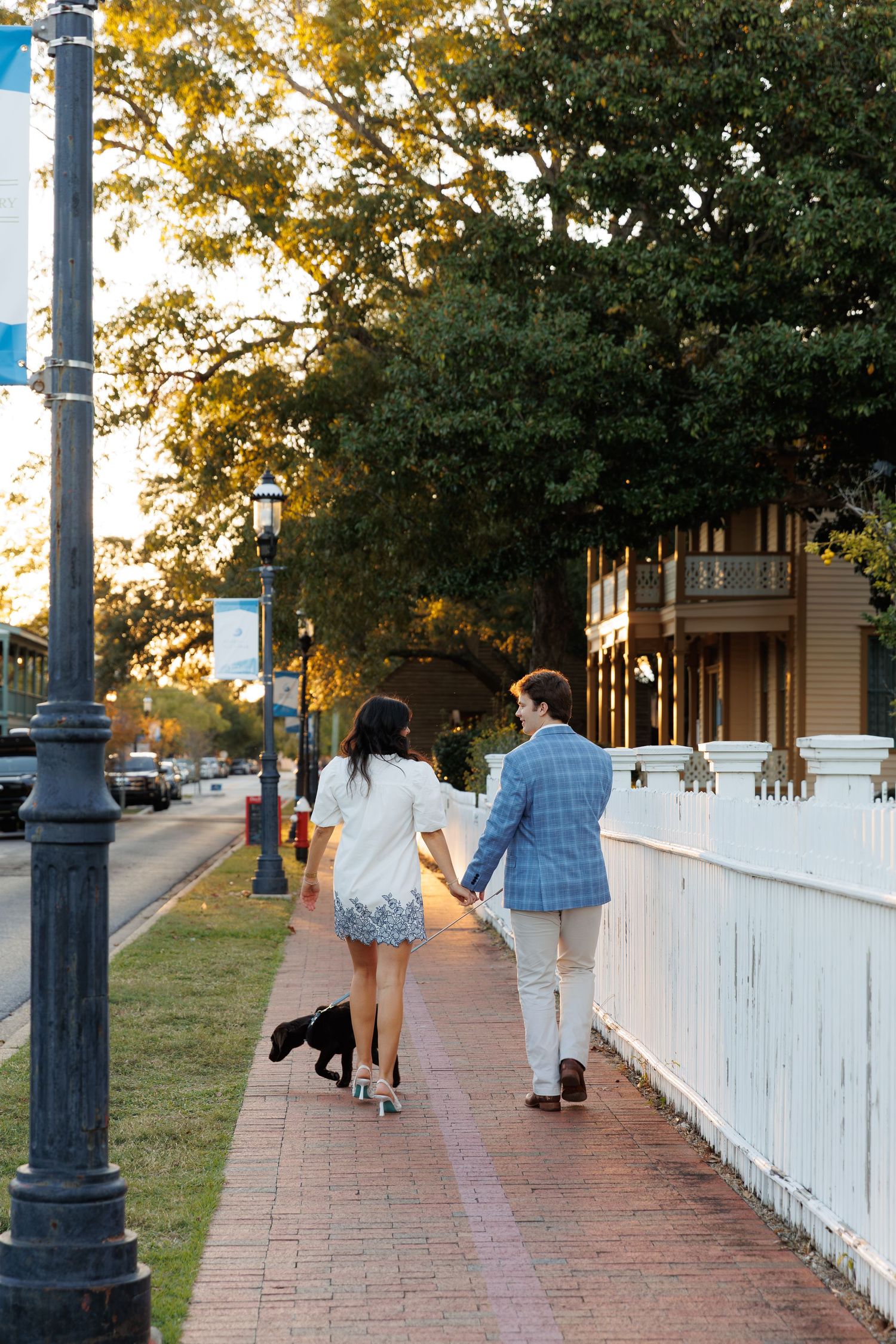 People walking along a white picket fence walkway at sunset with ornate lamp posts and autumn trees in background.
