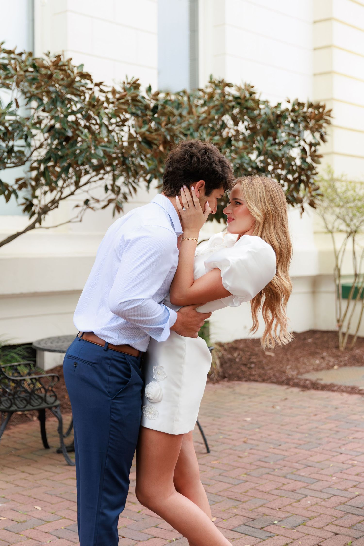 A couple shares an intimate moment on a brick patio outside a white building with decorative shrubs.