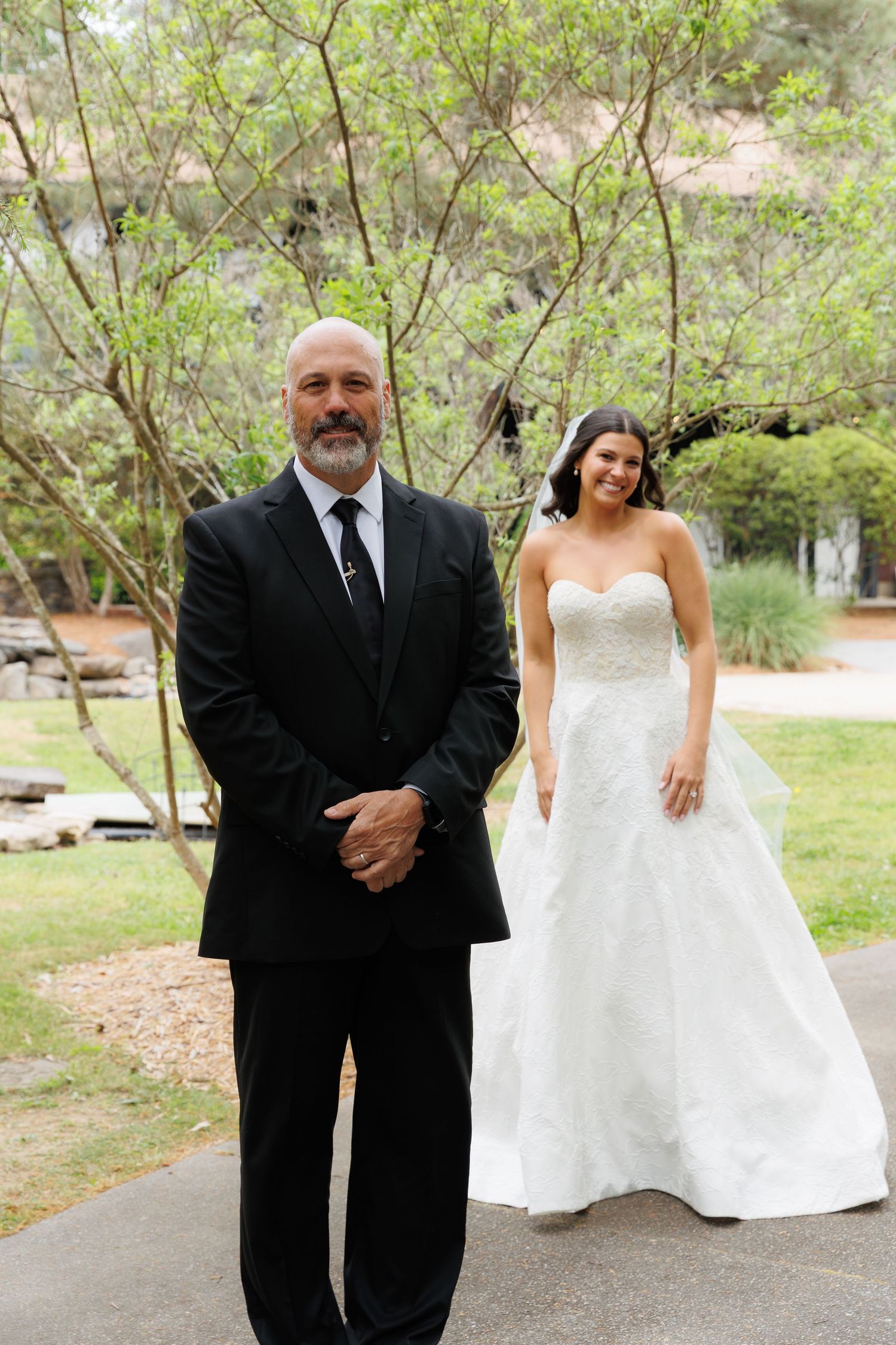 Bride in strapless white gown and veil stands behind a bald man in a black suit for a first look moment.