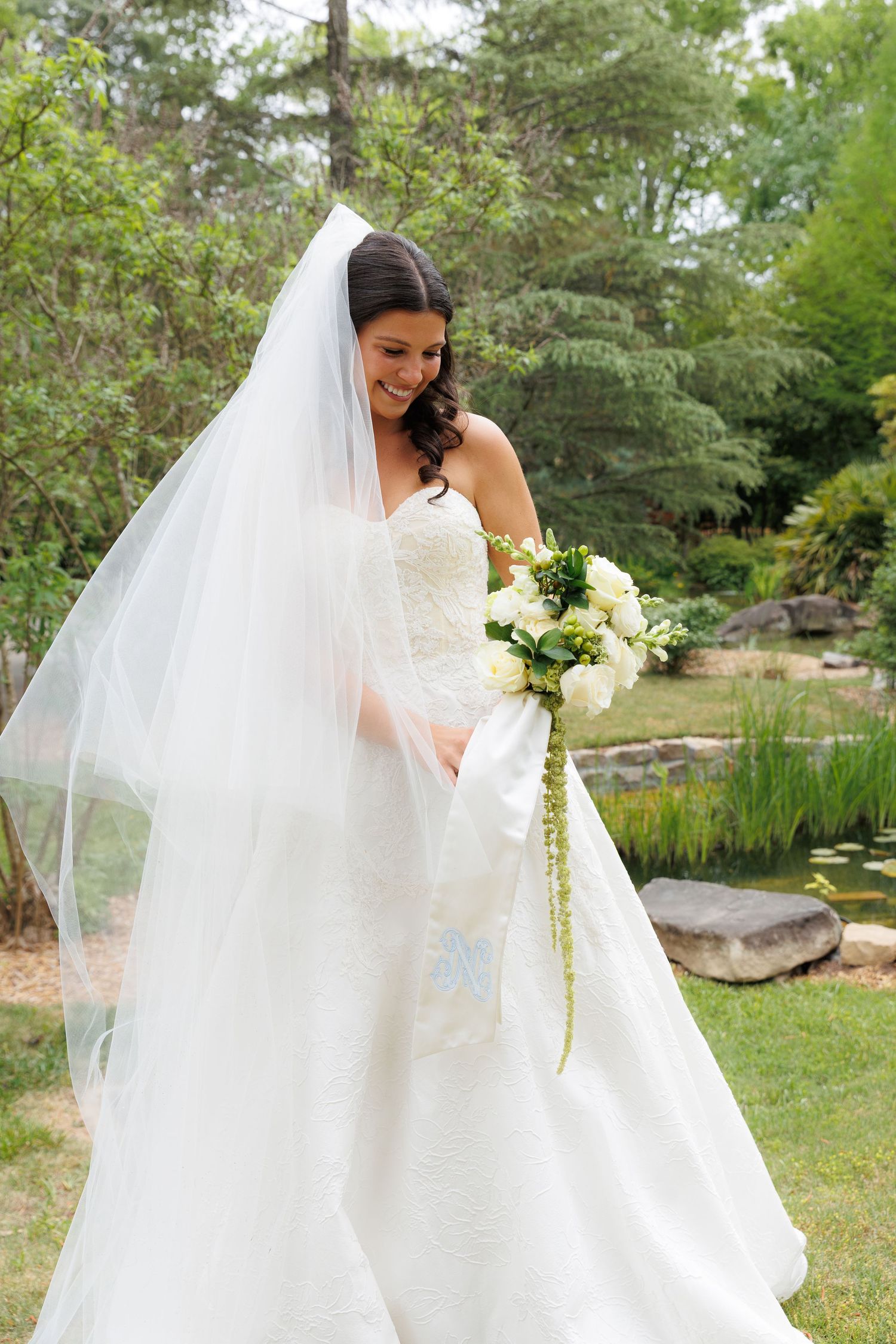 Bride in white strapless gown and flowing veil holds white bouquet outdoors in lush garden setting.