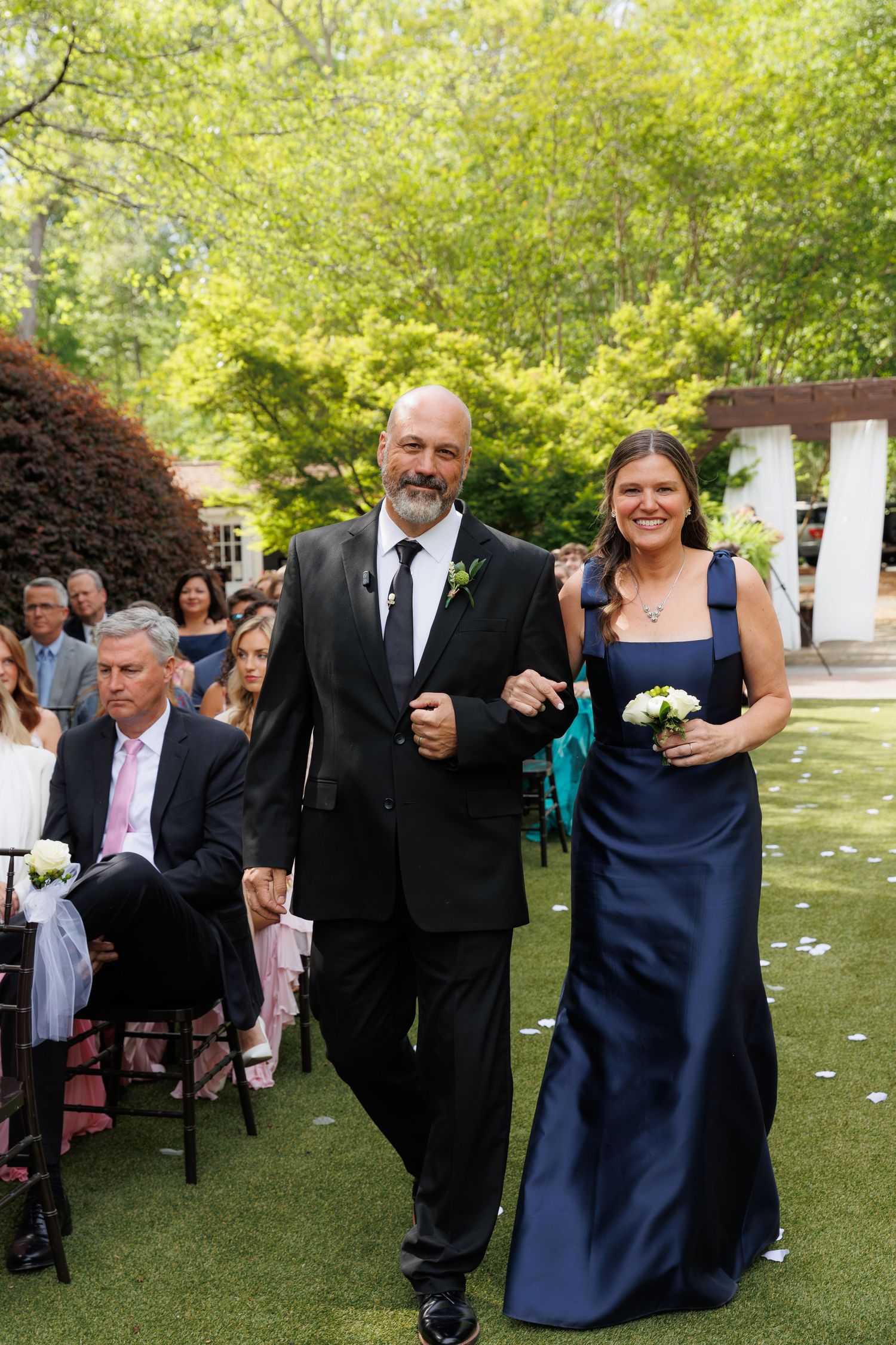 Elegant couple walking down outdoor wedding aisle, man in black tuxedo and woman in navy blue gown holding white flowers.