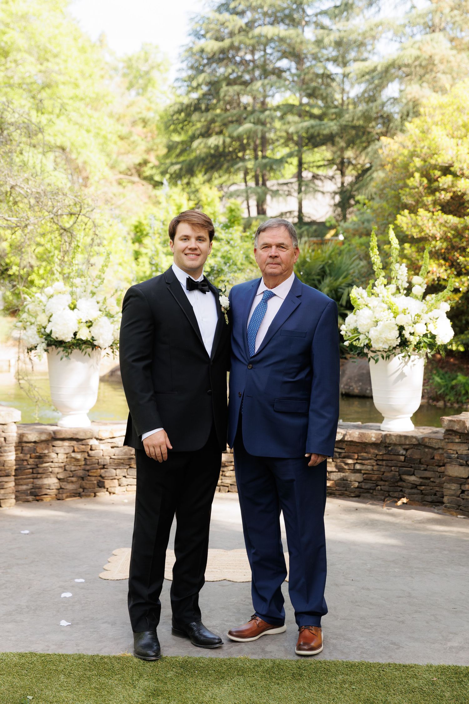 Two men in formal suits pose outdoors at a wedding ceremony with white floral arrangements.
