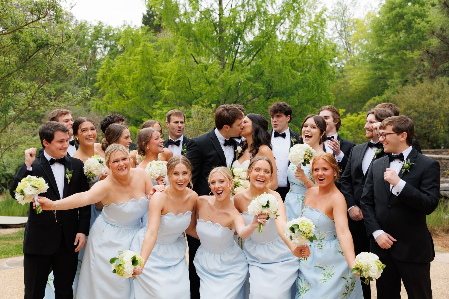 Wedding party poses outdoors, bridesmaids in light blue gowns and groomsmen in tuxedos, as bride and groom share a kiss.
