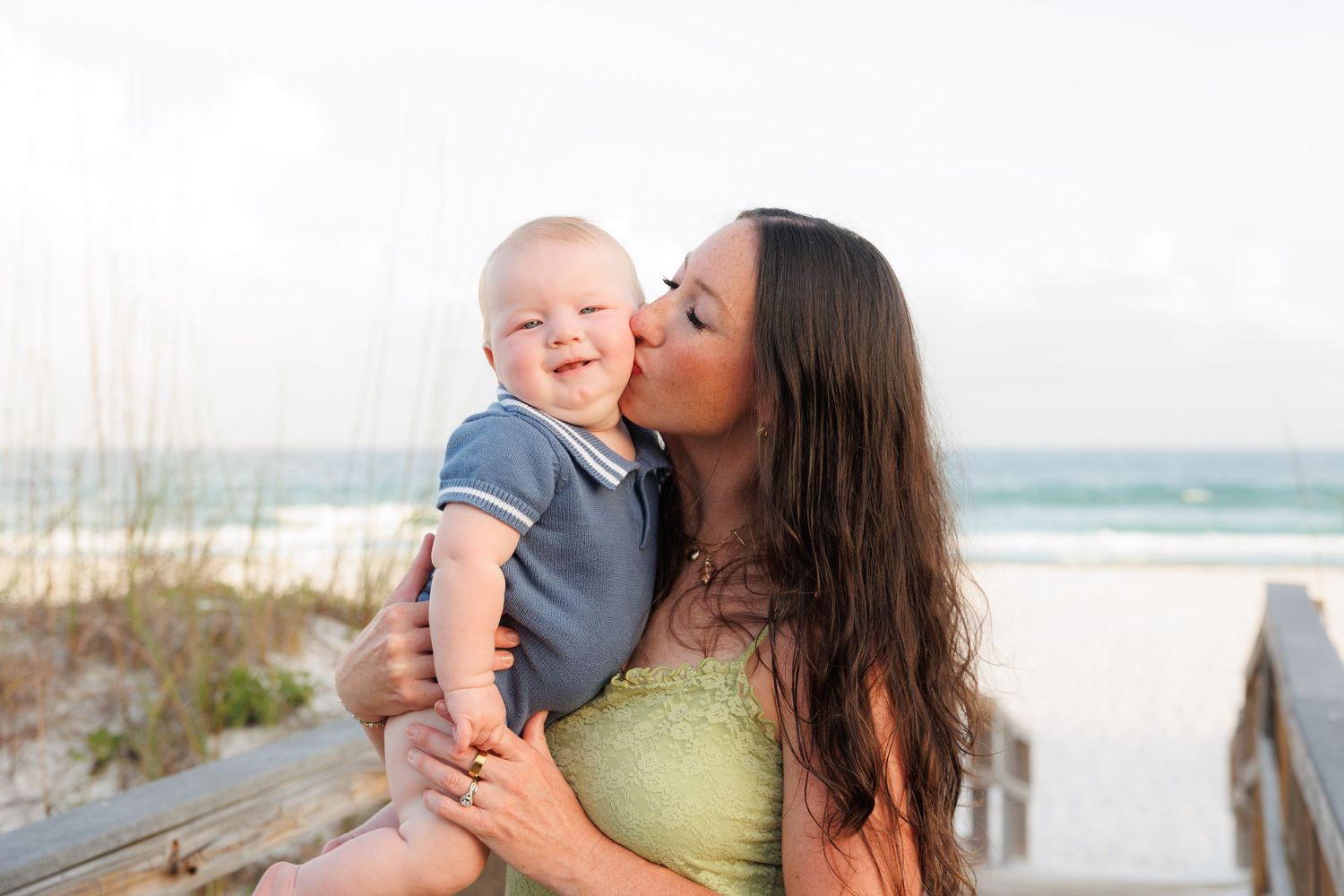 A mother kissing her smiling baby boy on a sunny beach boardwalk.