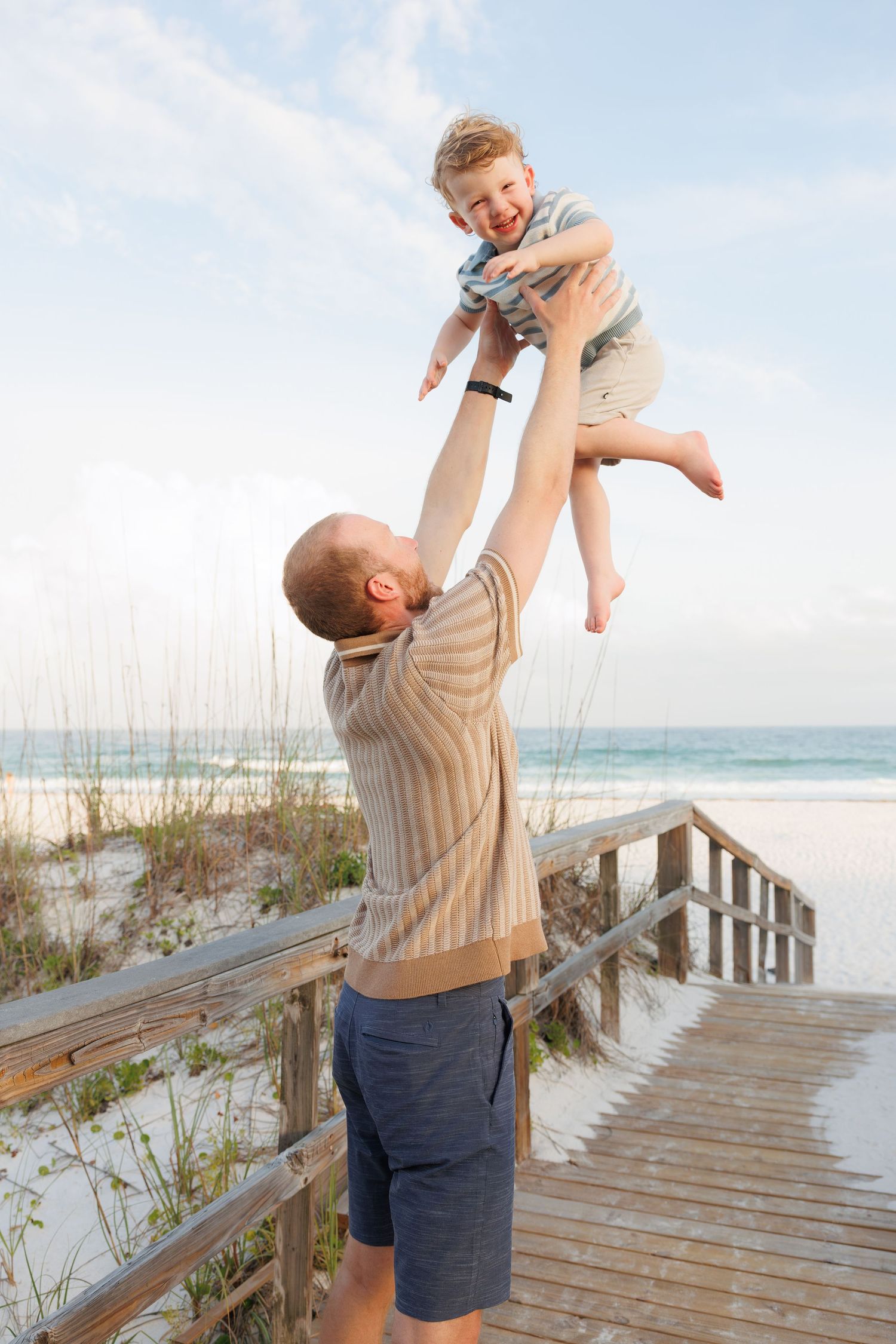 Father joyfully lifting laughing child high in the air on a sunny beach boardwalk with ocean in background.