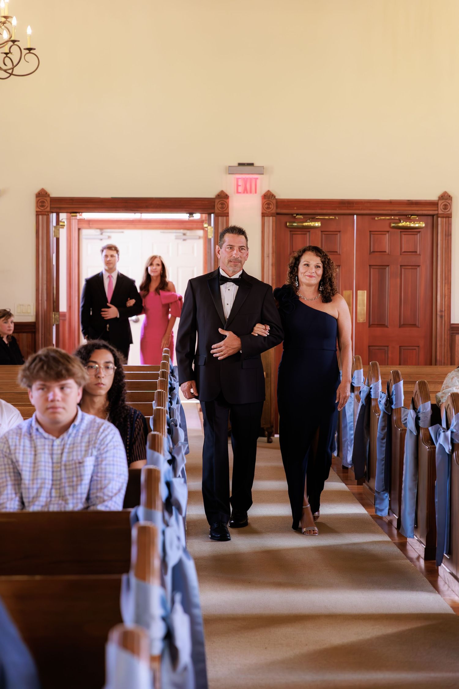 Church wedding guests sit in wooden pews decorated with light blue bows during ceremony preparations.