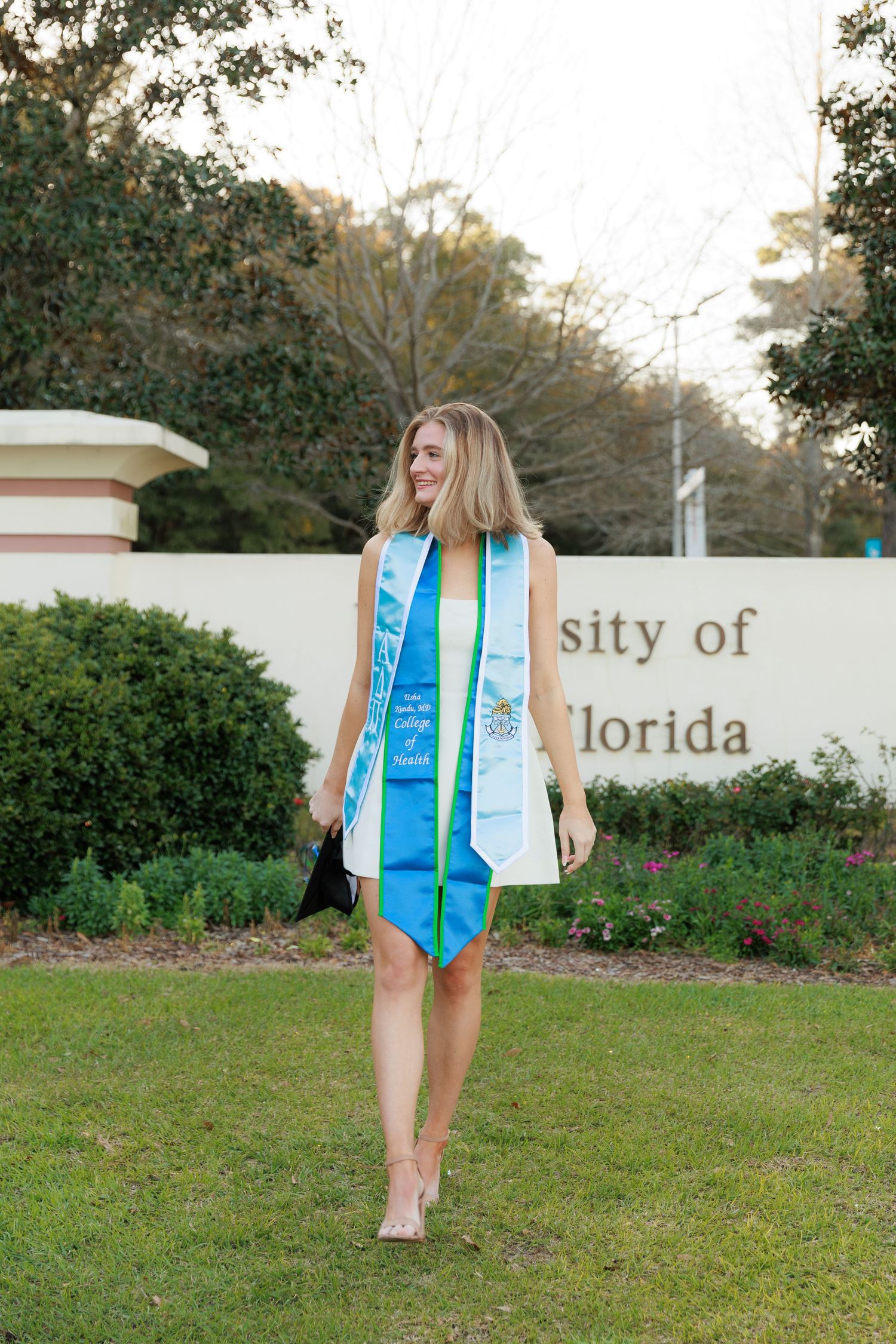 Graduate in blue dress and white graduation stole stands on lawn in front of University of Florida sign.